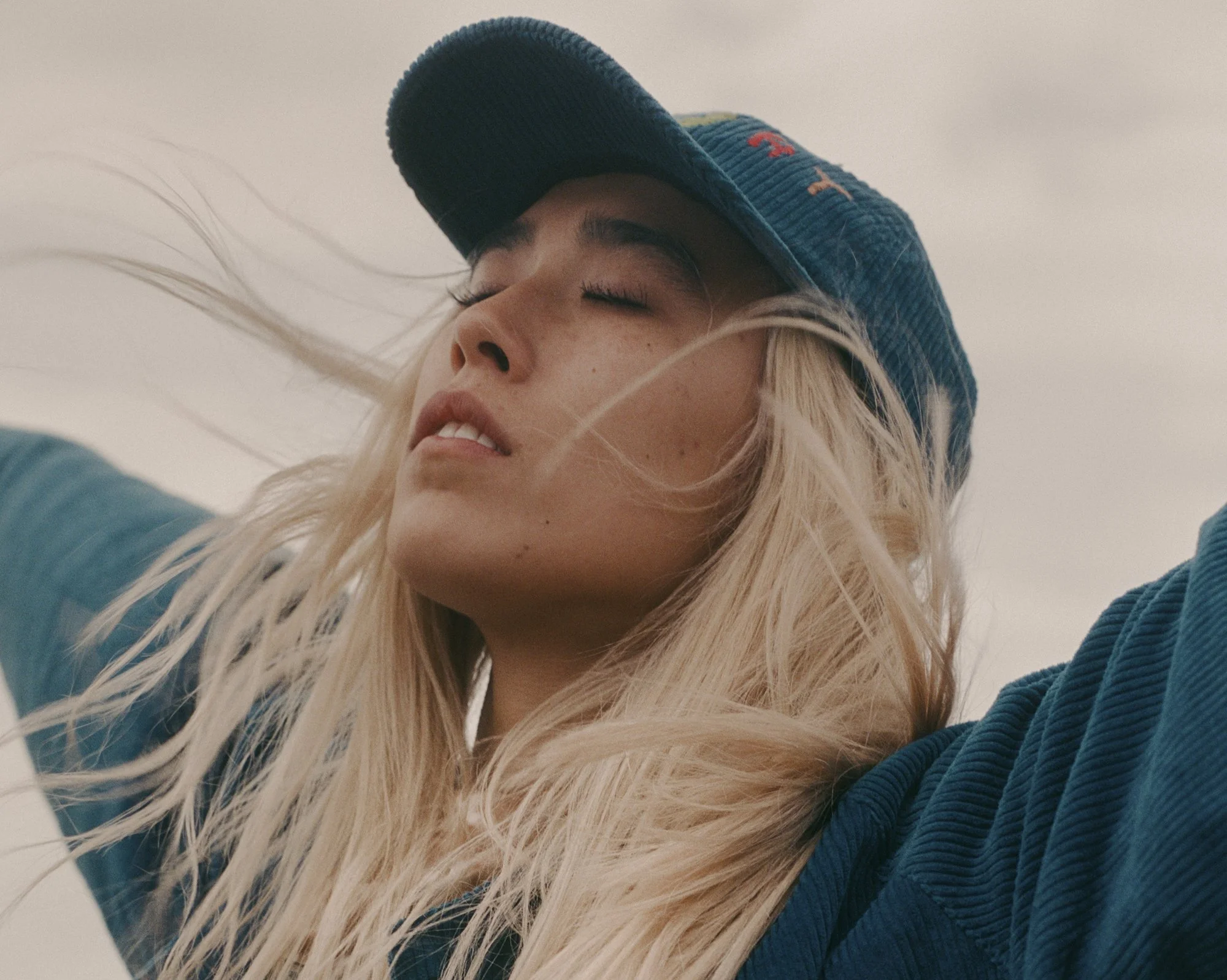 Close-up of a woman with long blonde hair, wearing a blue cap and jacket, with eyes closed and hair blowing in the wind.