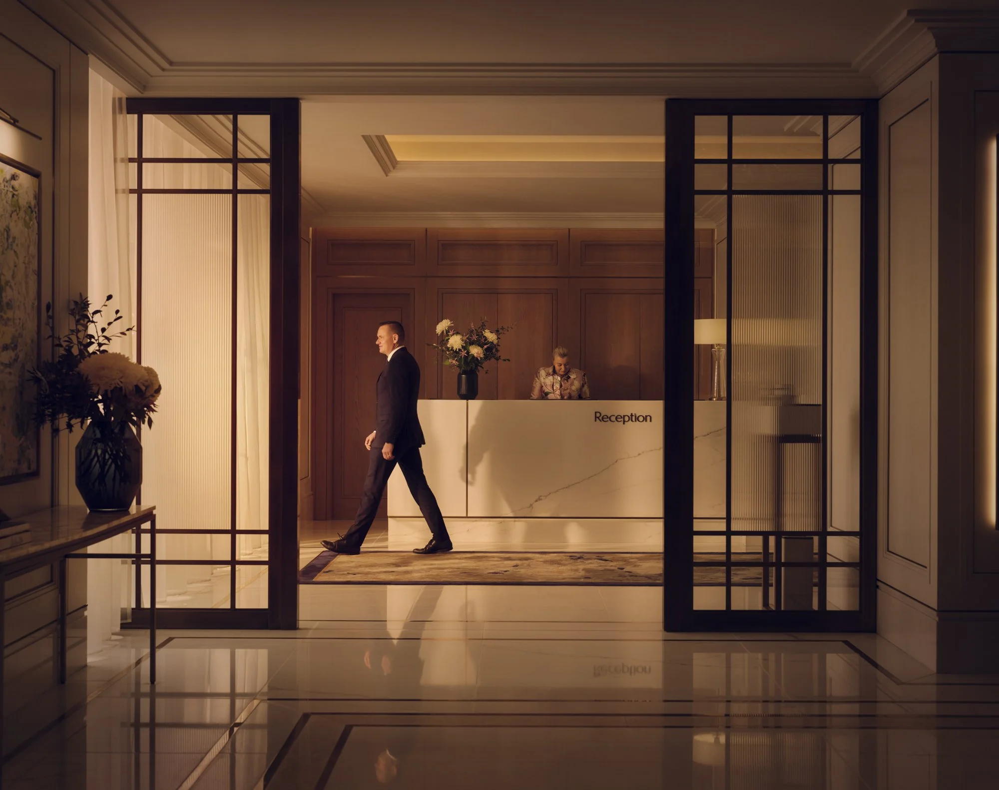 A man in a black suit walking in front of a reception desk in a hotel lobby.