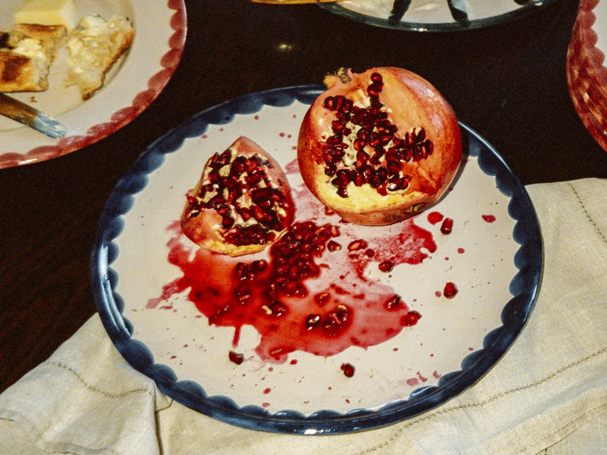 A plate with an opened pomegranate, pomegranate seeds spilled on the plate, and some juice. Part of another plate with some food remains is visible in the background.