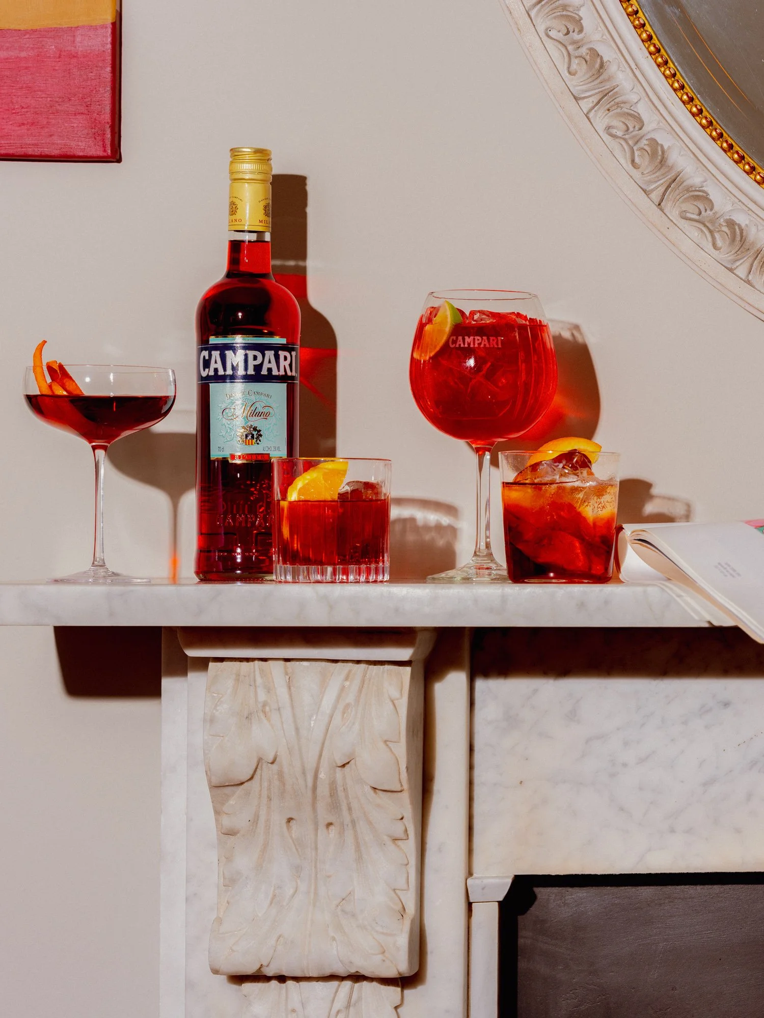 A bottle of Campari and four glasses filled with red cocktails, garnished with citrus slices, on a white marble table.