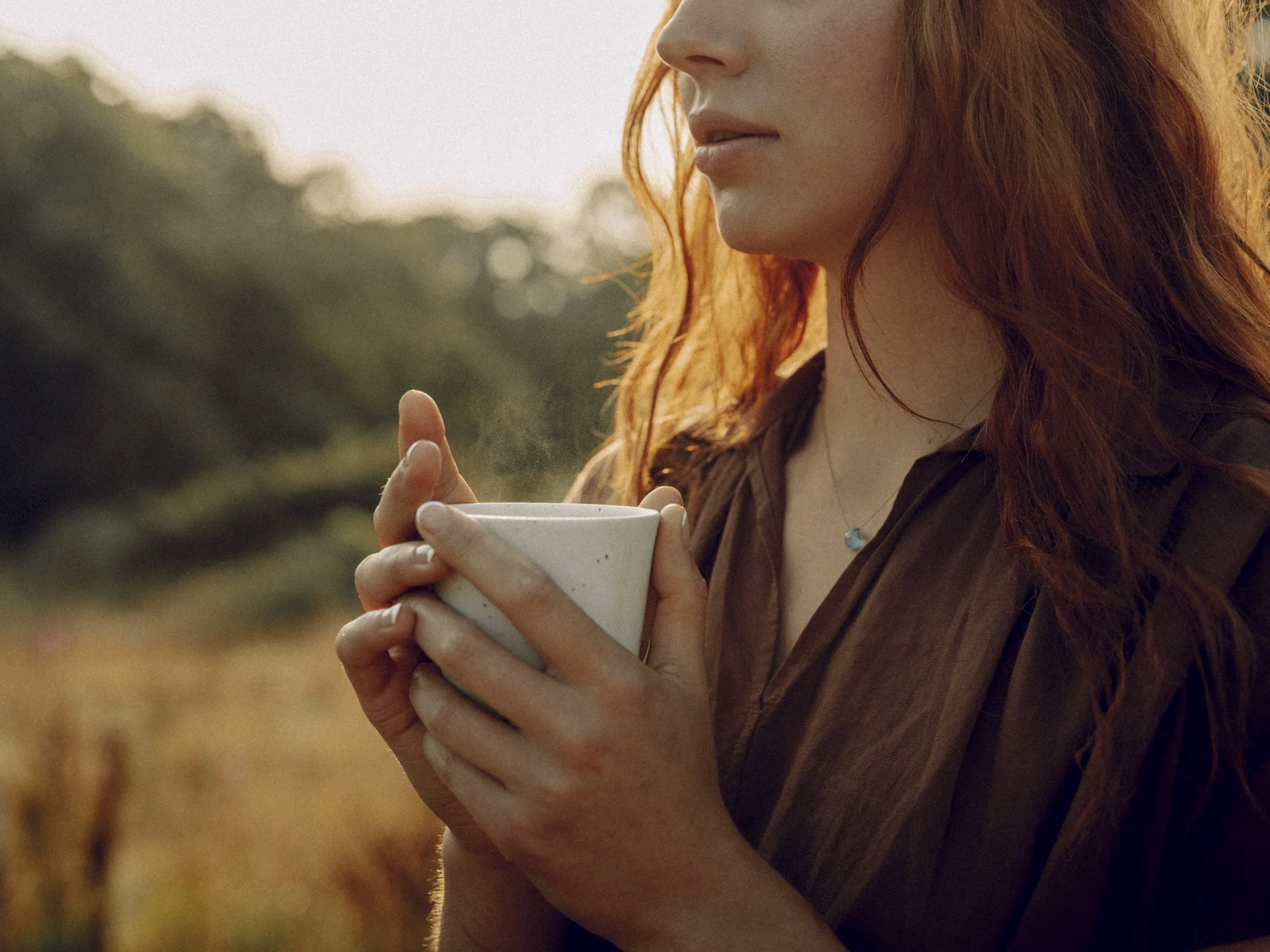 A woman holding a steaming mug outdoors in a field during the golden hour.