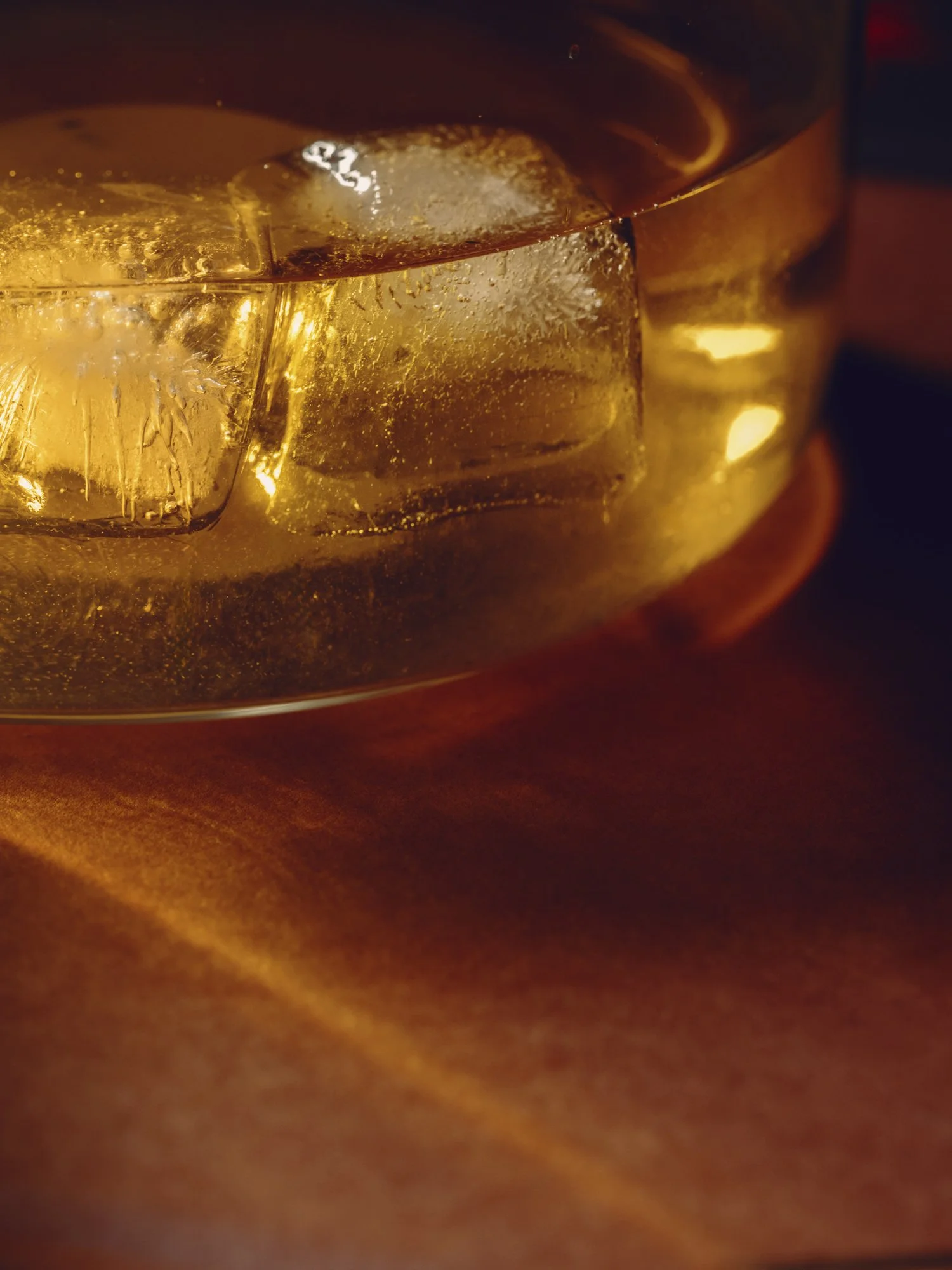 Close-up of a glass of whiskey with ice cubes, viewed in warm, ambient light.