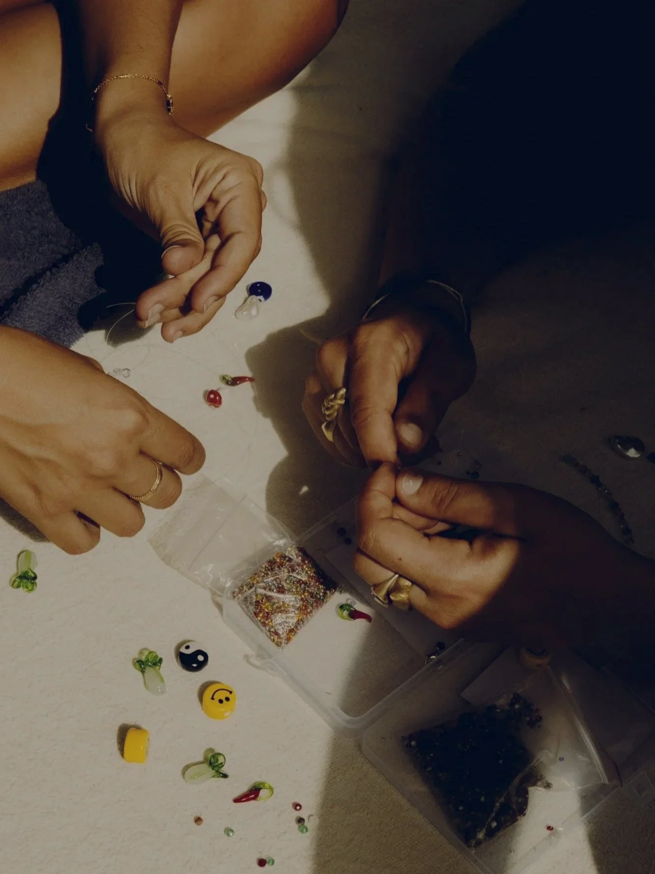 Two people are making jewelry or crafts with beads and other small decorative items on a white surface.