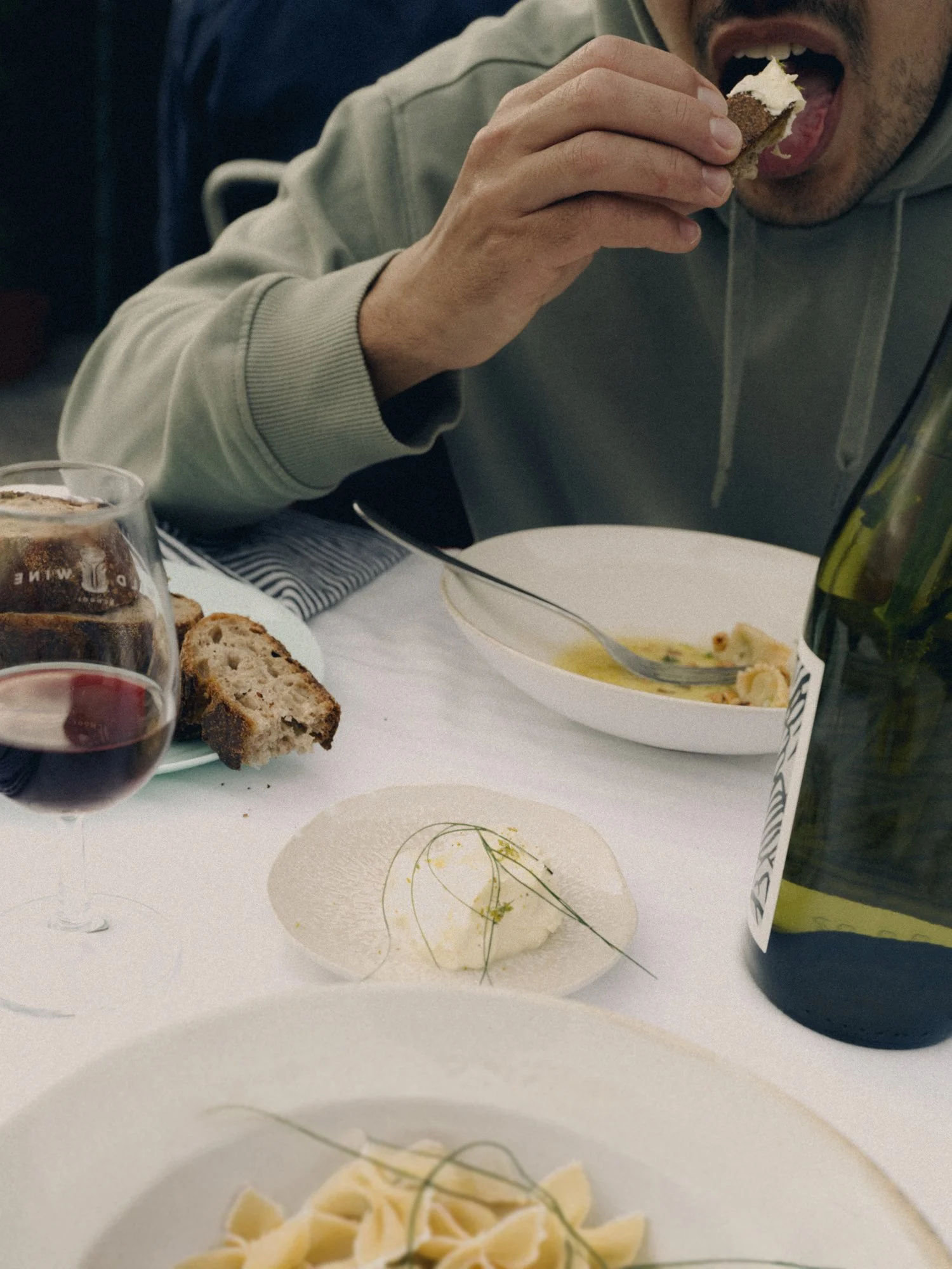 Person sitting at a table eating bread with butter, with a glass of red wine, plates of pasta, and a bottle of wine on the table.