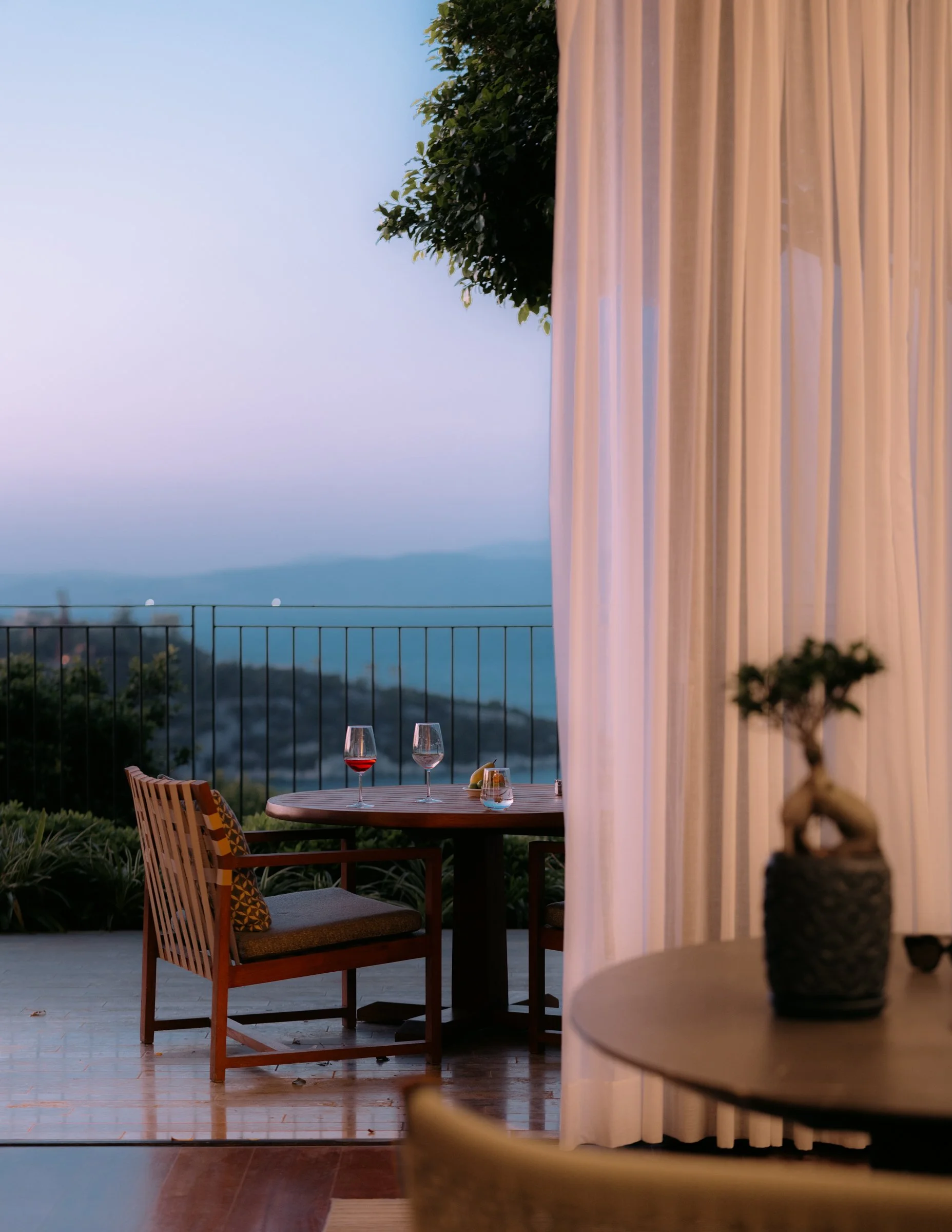 A cozy outdoor balcony scene during dusk with a round wooden table set with two glasses of red wine, a glass of water, and a lemon slice. Two wooden chairs with cushions are arranged around the table. The balcony is surrounded by a railing with a sce