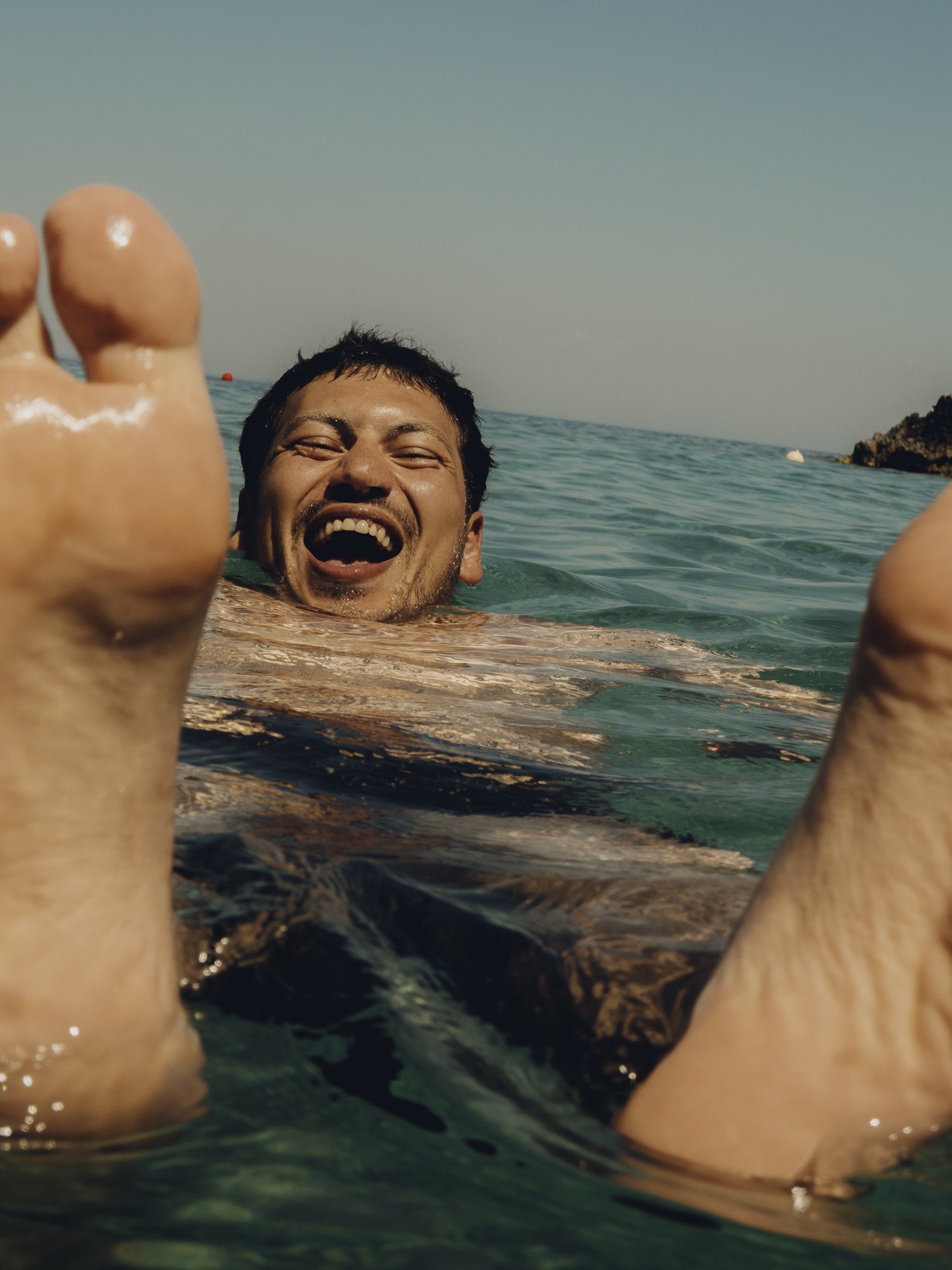 Man laughing and smiling while floating in the ocean with arms raised, clear sky, and distant shoreline.