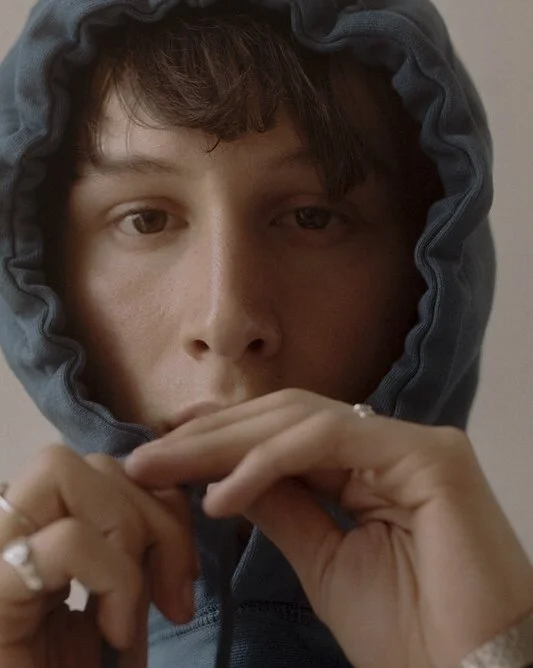 Close-up of a person wearing a hoodie, looking directly at the camera, with their hands raised to their face.