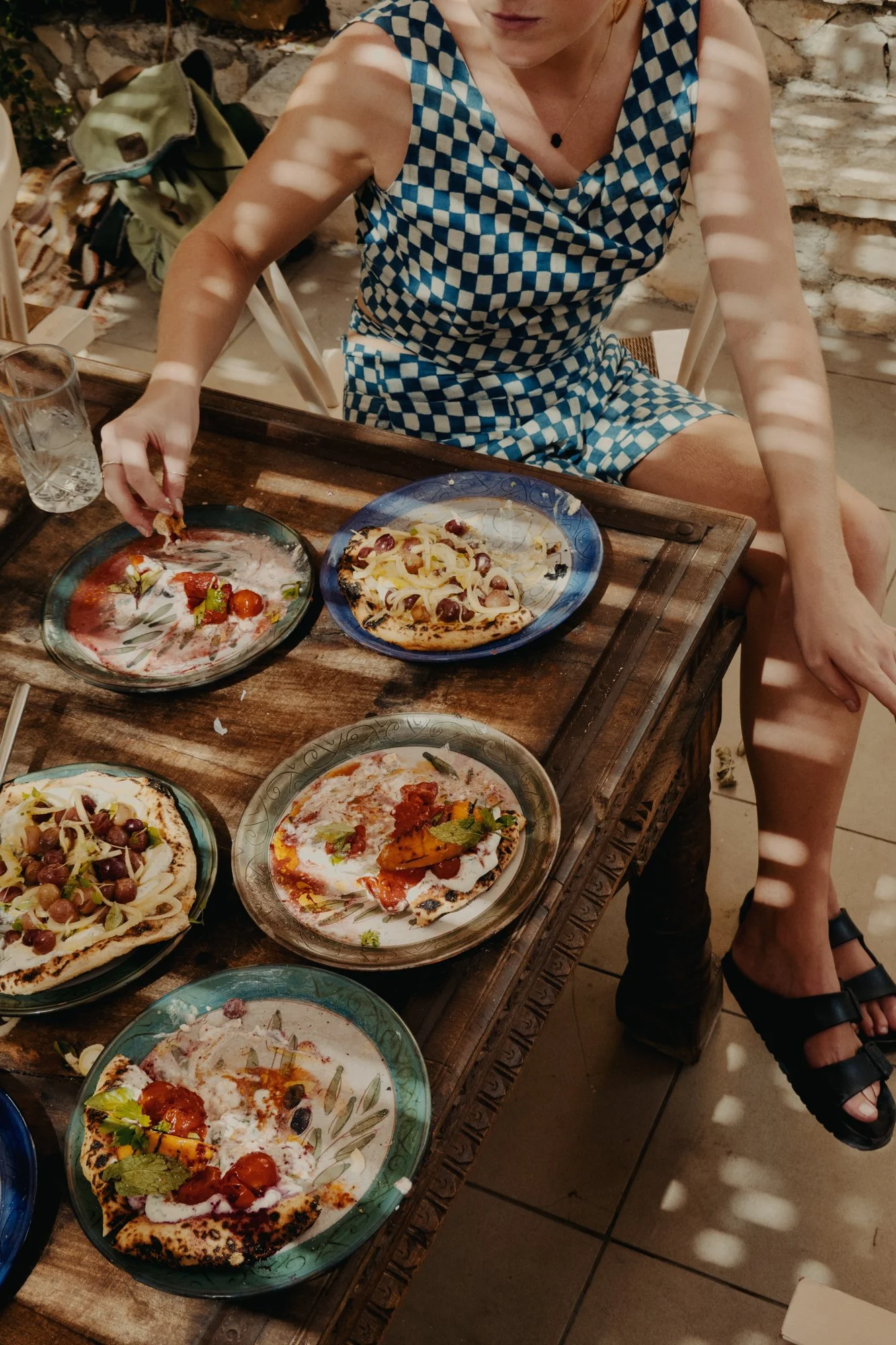 A person sitting at a wooden table with several plates of pizza, wearing a blue and white checkered sleeveless dress, with sunlight filtering through shadows cast by nearby blinds.