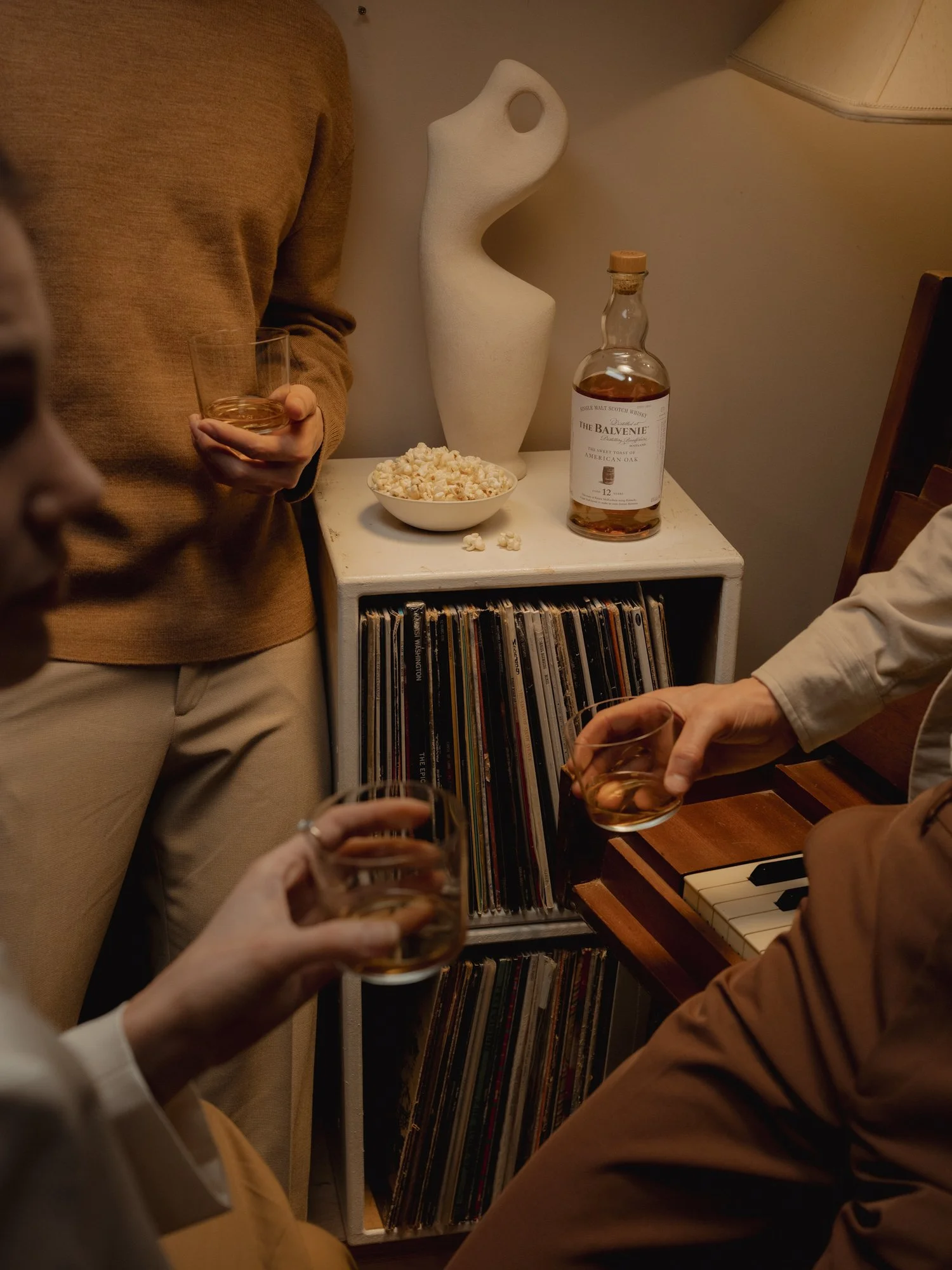 People having a gathering, holding glasses of whiskey, in a cozy room with a vinyl record collection, popcorn bowl, and bottle of whiskey on a small white table, along with a decorative sculpture and a lamp.