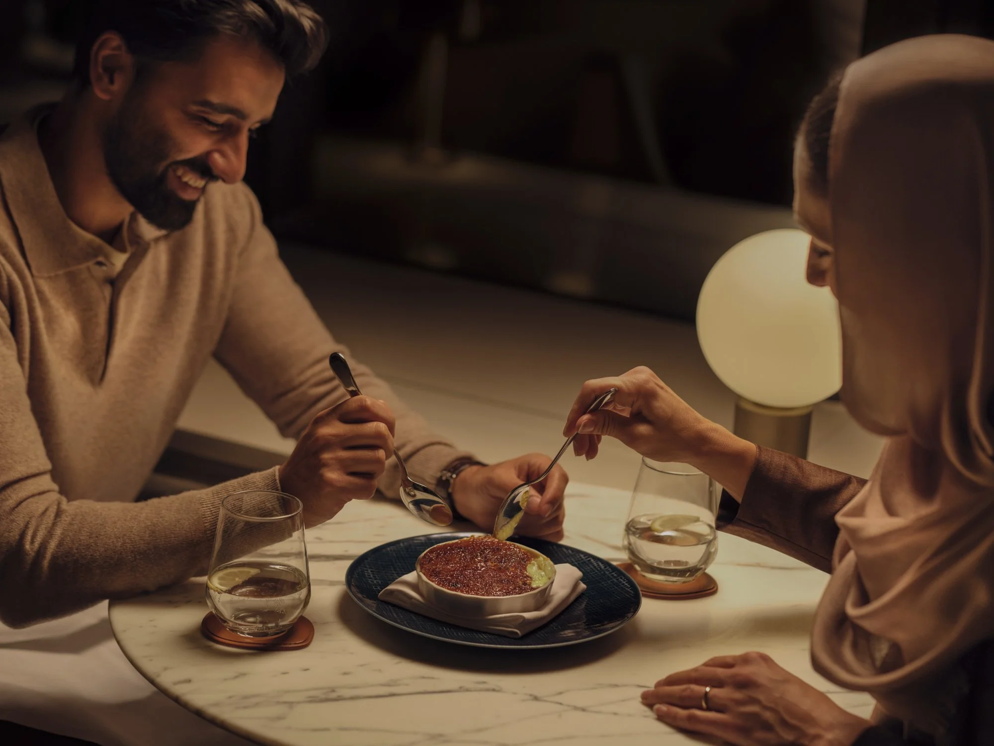 A man and woman sharing dessert at a cozy restaurant table, smiling and lifting spoons. The table has a round marble surface with two glasses of water, and a lit lamp provides warm lighting.
