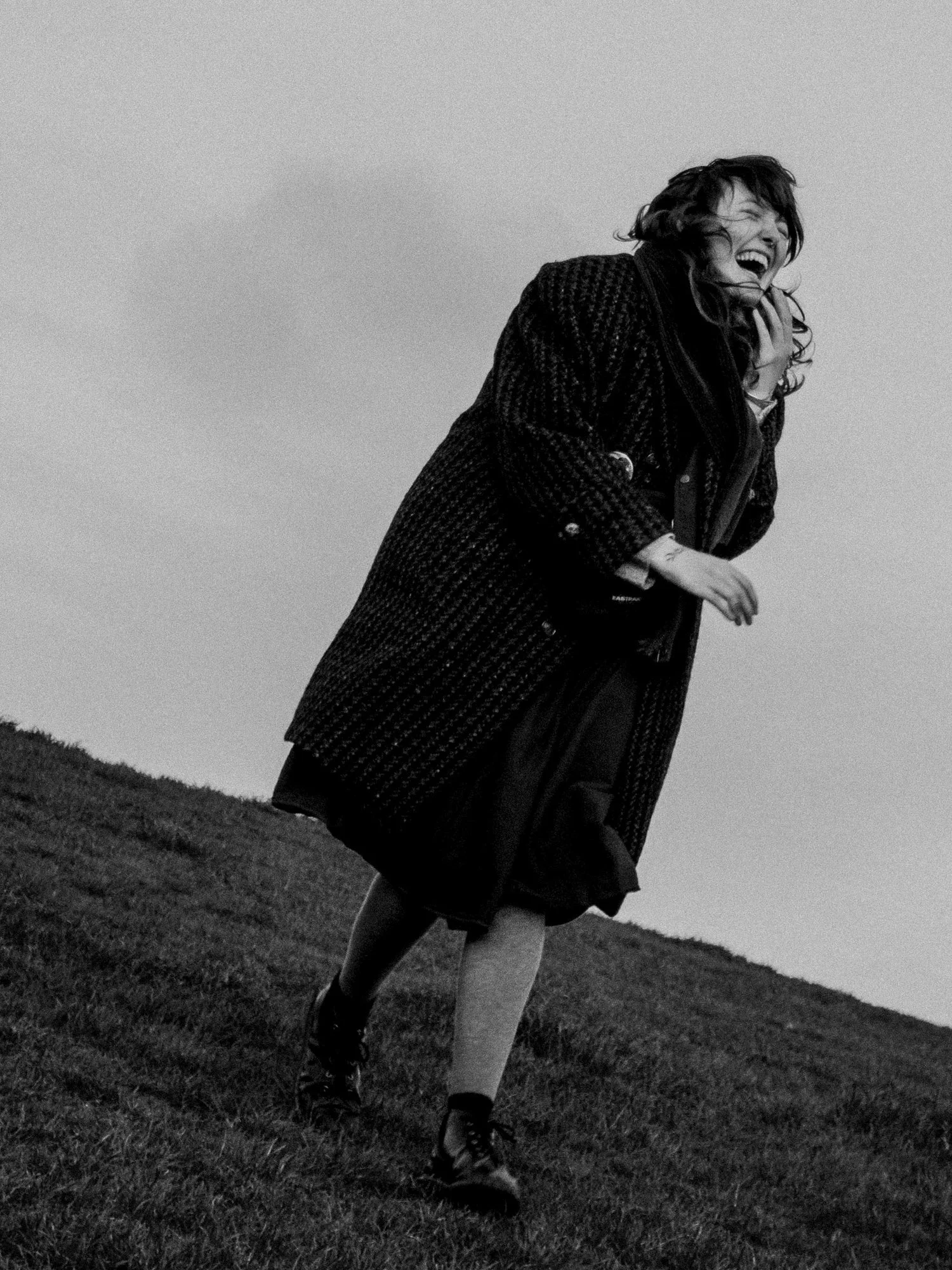 A woman laughing and leaning forward on a grassy field, wearing a dark coat and shoes, with hair blowing in the wind.