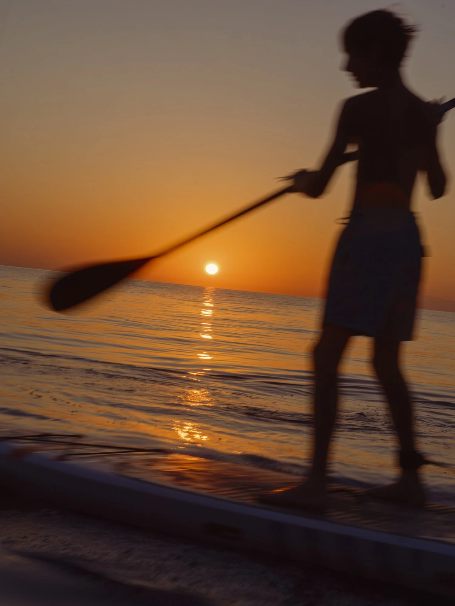 Silhouette of a woman paddleboarding on the ocean at sunset, with the sun low on the horizon and casting a warm glow over the water.