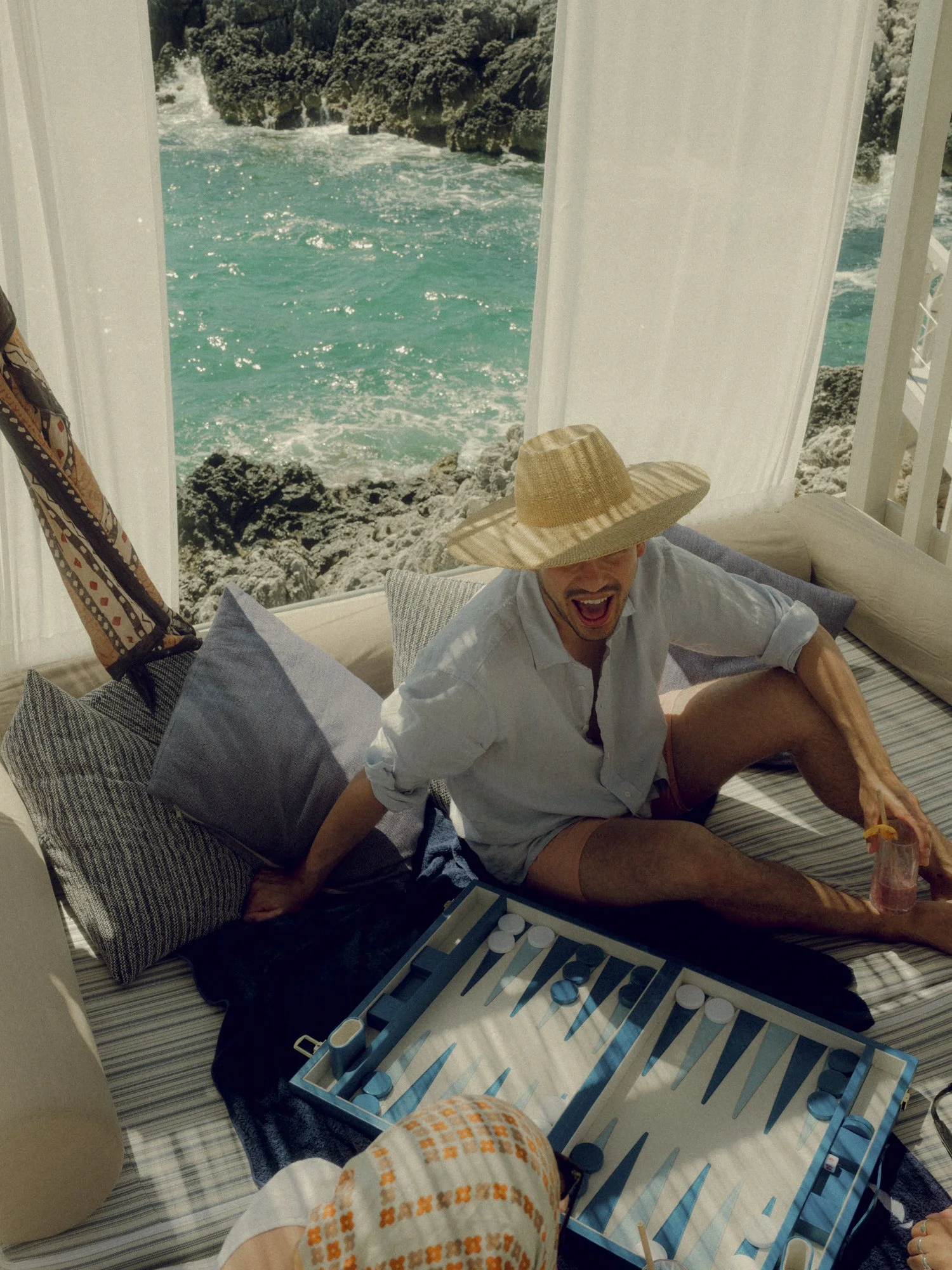 A man wearing a large straw hat, white shirt, and shorts is sitting on a bed while playing a game of backgammon. He is holding a drink with a small umbrella and appears to be laughing or celebrating. The background shows a window with a view of the o