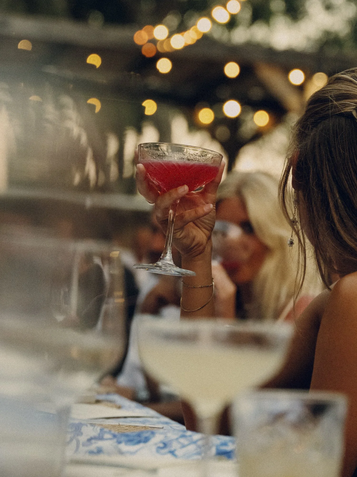 Person holding a red cocktail glass at a social gathering, with warm string lights and blurred people in the background.