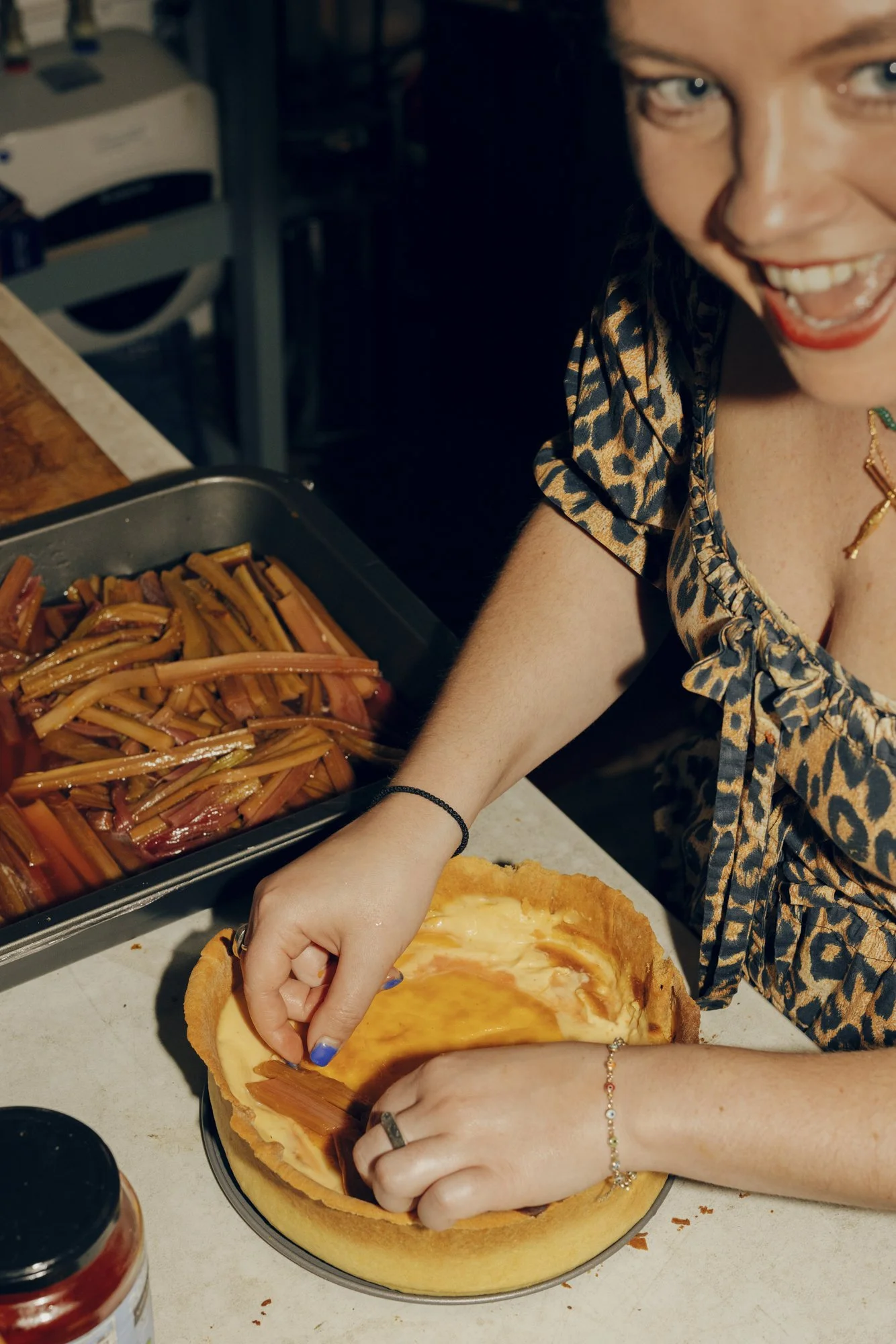 A woman preparing a rhubarb pie in a kitchen, with a baking crust in the pan and a tray of rhubarb stalks on the counter.