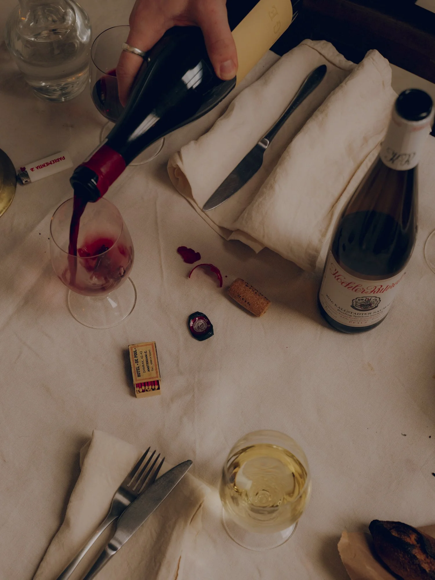 A table with a glass of white wine, a glass of red wine, a bottle of red wine being poured into a glass, and various tableware including a fork, knife, and napkin. There are also packages of matches, a cork, and a rolled-up napkin.