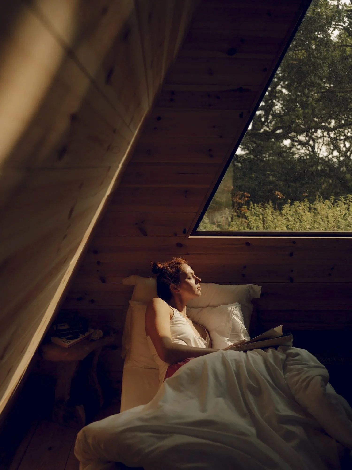 A woman lies in bed with her eyes closed, reading a book, in a room with wooden walls and a large skylight window showing trees outside.