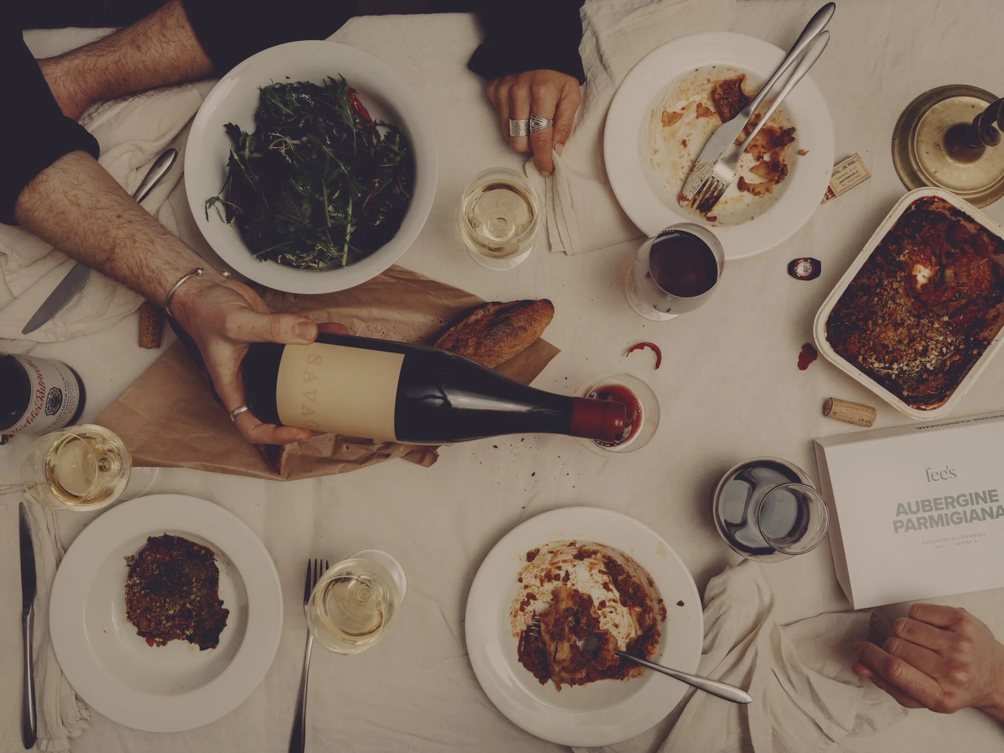 A dining table with leftover food, glasses of wine and water, a bottle of wine, and some plates of partially eaten dishes, with people’s hands and arms visible.