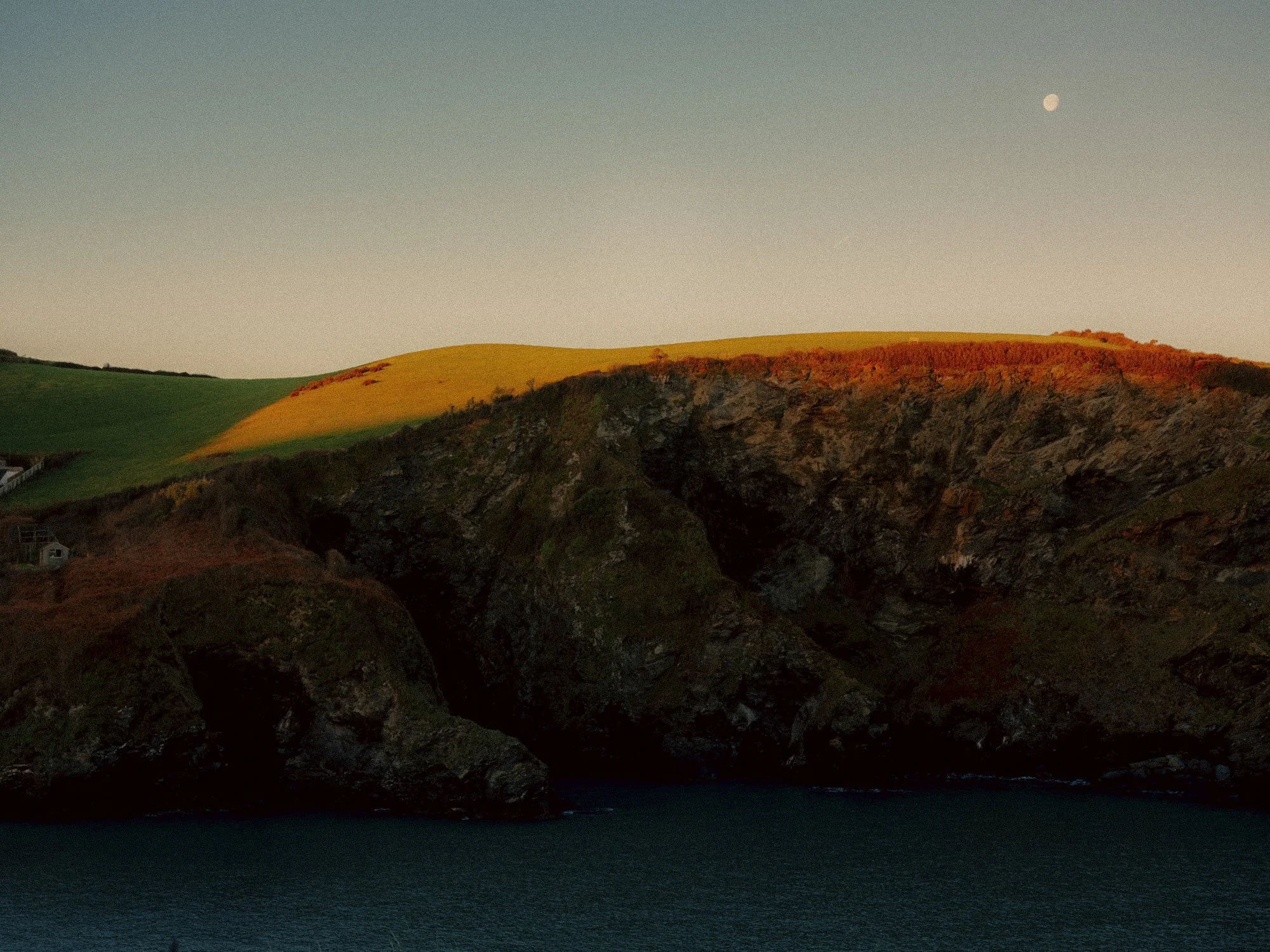 Hilly landscape with green and brown grass, rocky hillside, and a body of water in the foreground. The sky is clear with a visible moon.