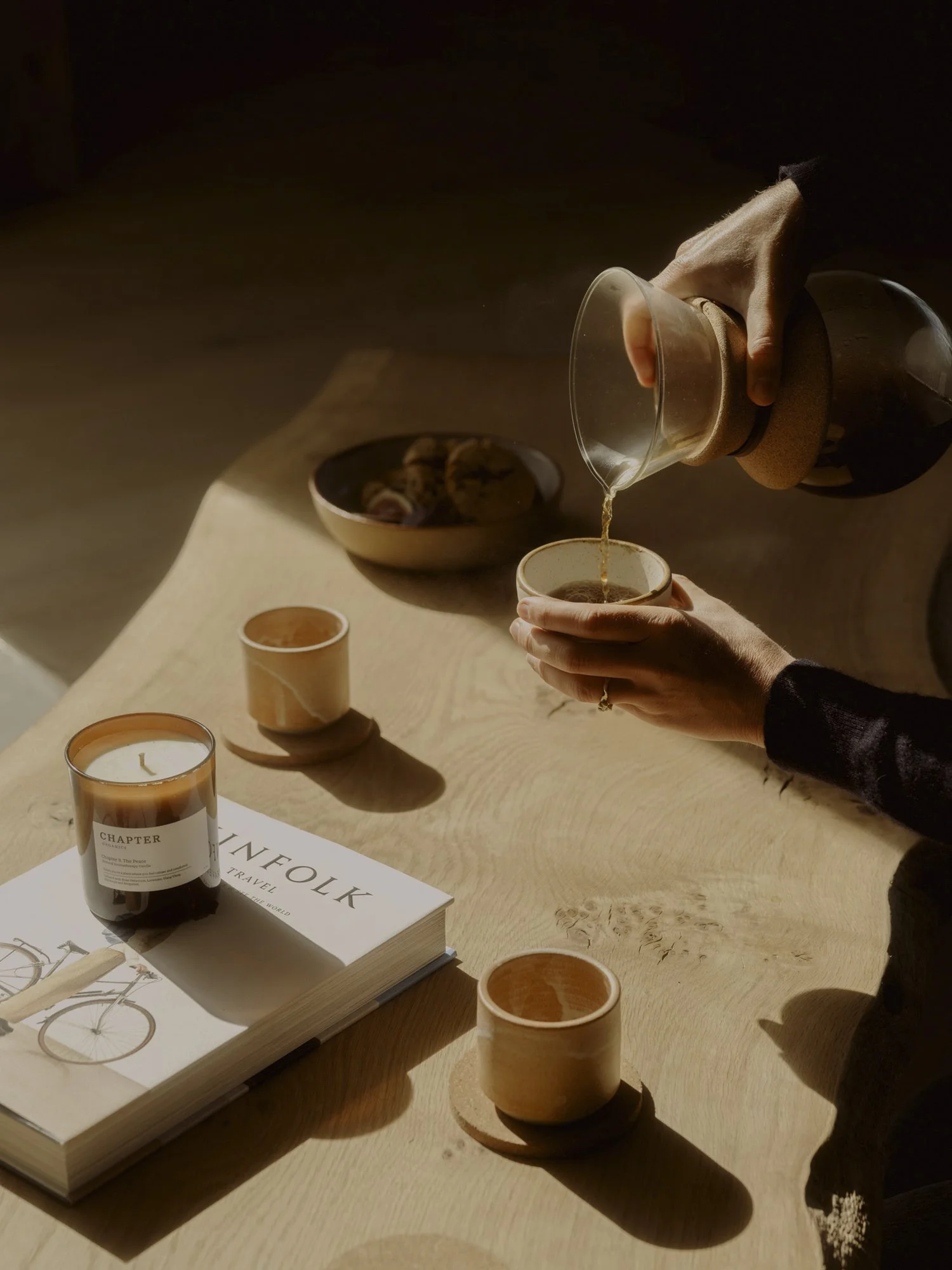 Person pouring coffee into a cup on a wooden table with a magazine, candles, and bowls.