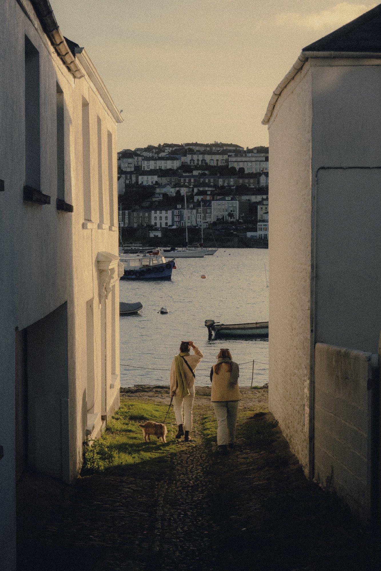 Two women walking a dog through a narrow alley towards a harbor with boats at sunset.
