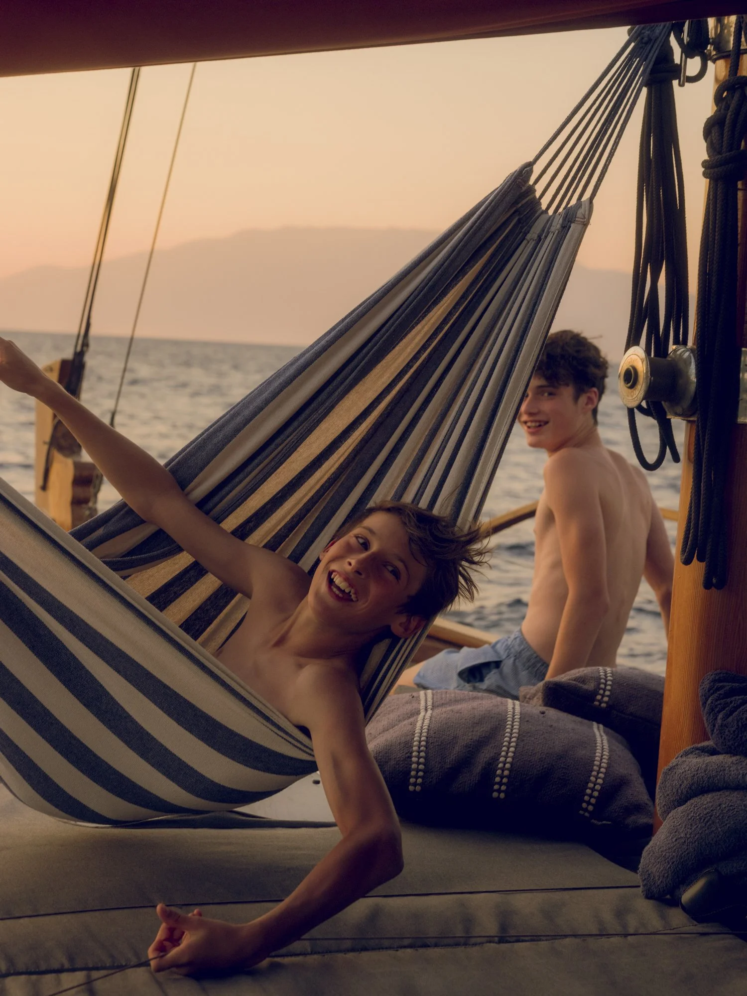 Two boys enjoying on a boat with a hammock during sunset, one lying in the hammock and smiling at the camera, the other standing nearby smiling, with the ocean and mountains in the background.