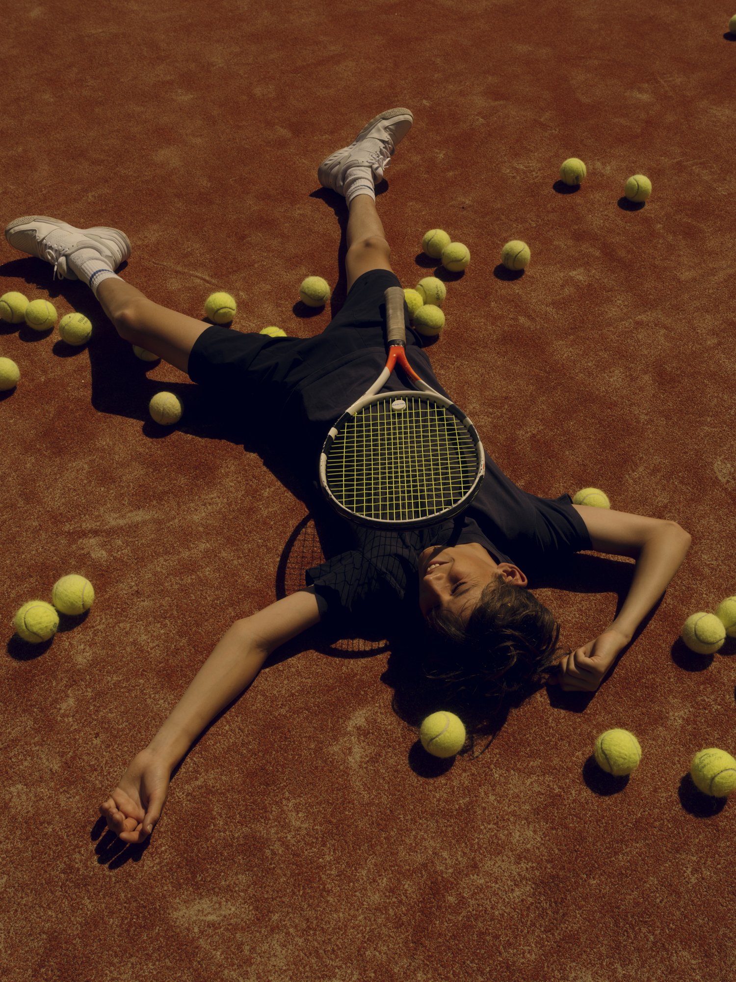 A person lying on a tennis court surrounded by tennis balls, holding a tennis racket on their stomach, with their eyes closed.