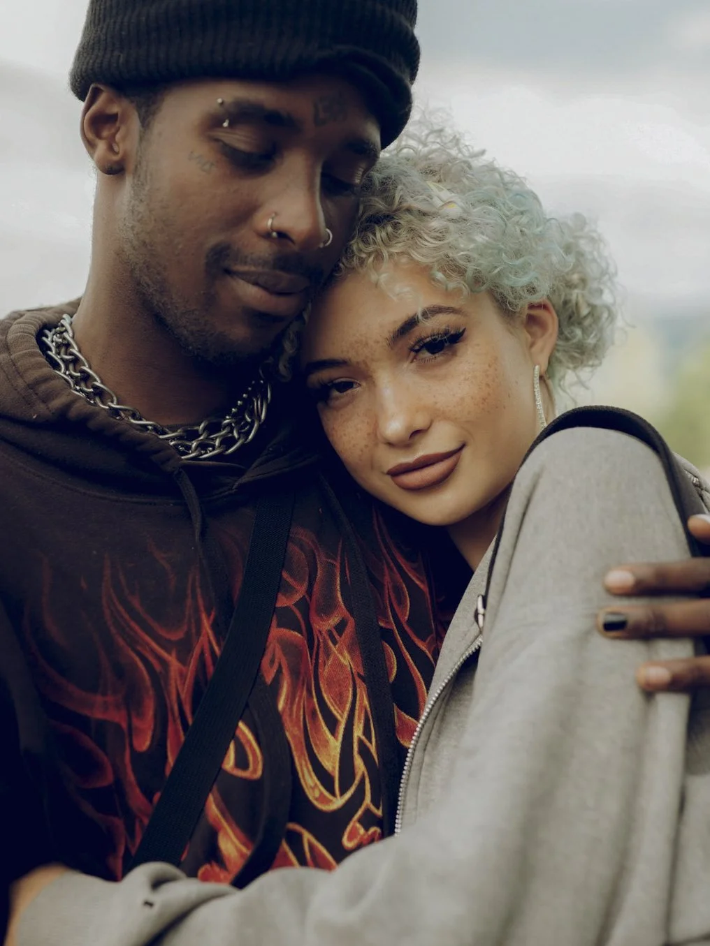 A close-up of a couple taking a selfie outdoors. The man has dark skin, multiple facial piercings, wearing a beanie, chains, and a dark shirt with red design. The woman has fair skin, curly platinum blonde hair, freckles, and is wearing makeup and ea
