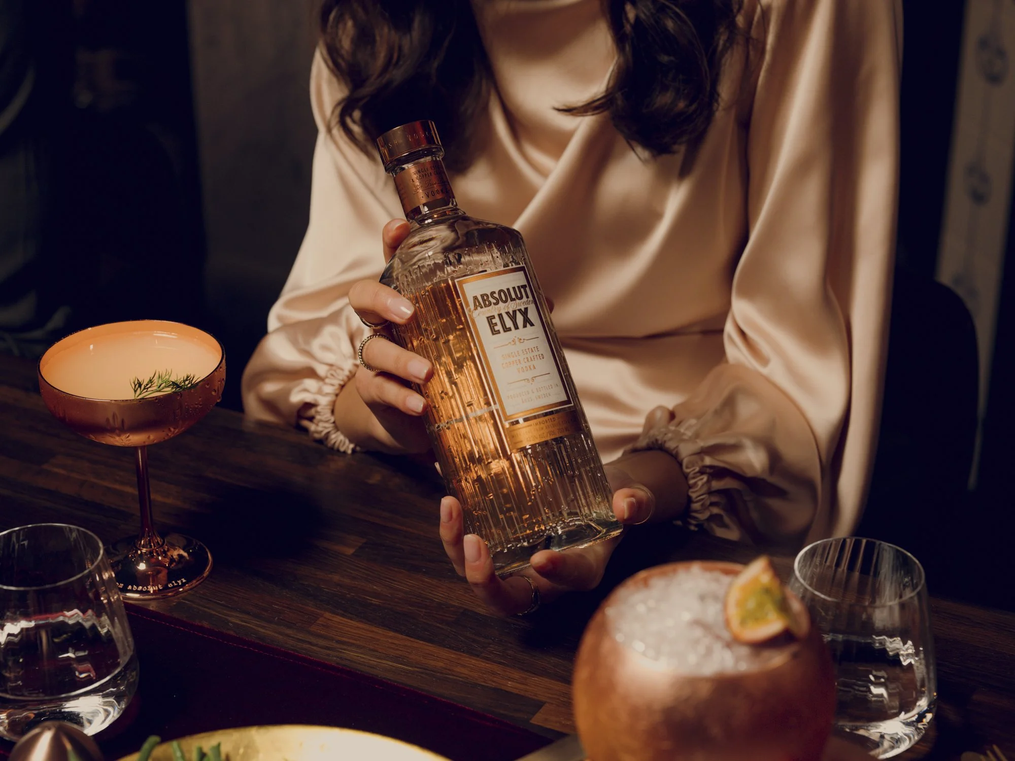 A woman wearing a beige satin blouse holds a bottle of Absolut Elyx vodka at a bar with glasses and cocktails on the table.
