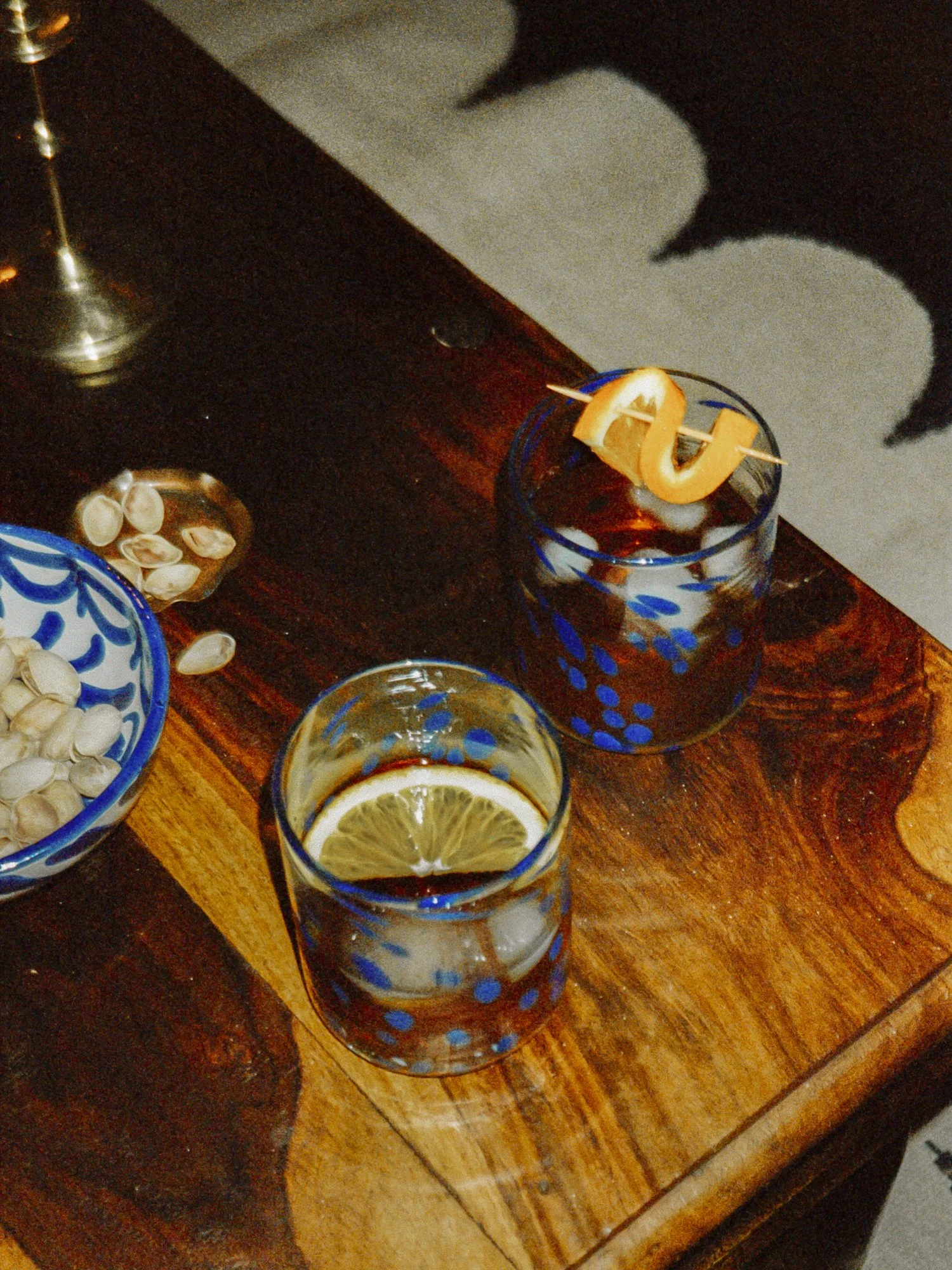 Two glasses of dark beverage with lemon slices, one garnished with a curly lemon peel and a skewer, on a wooden table. A bowl of pistachios and a silver candle holder are also visible.