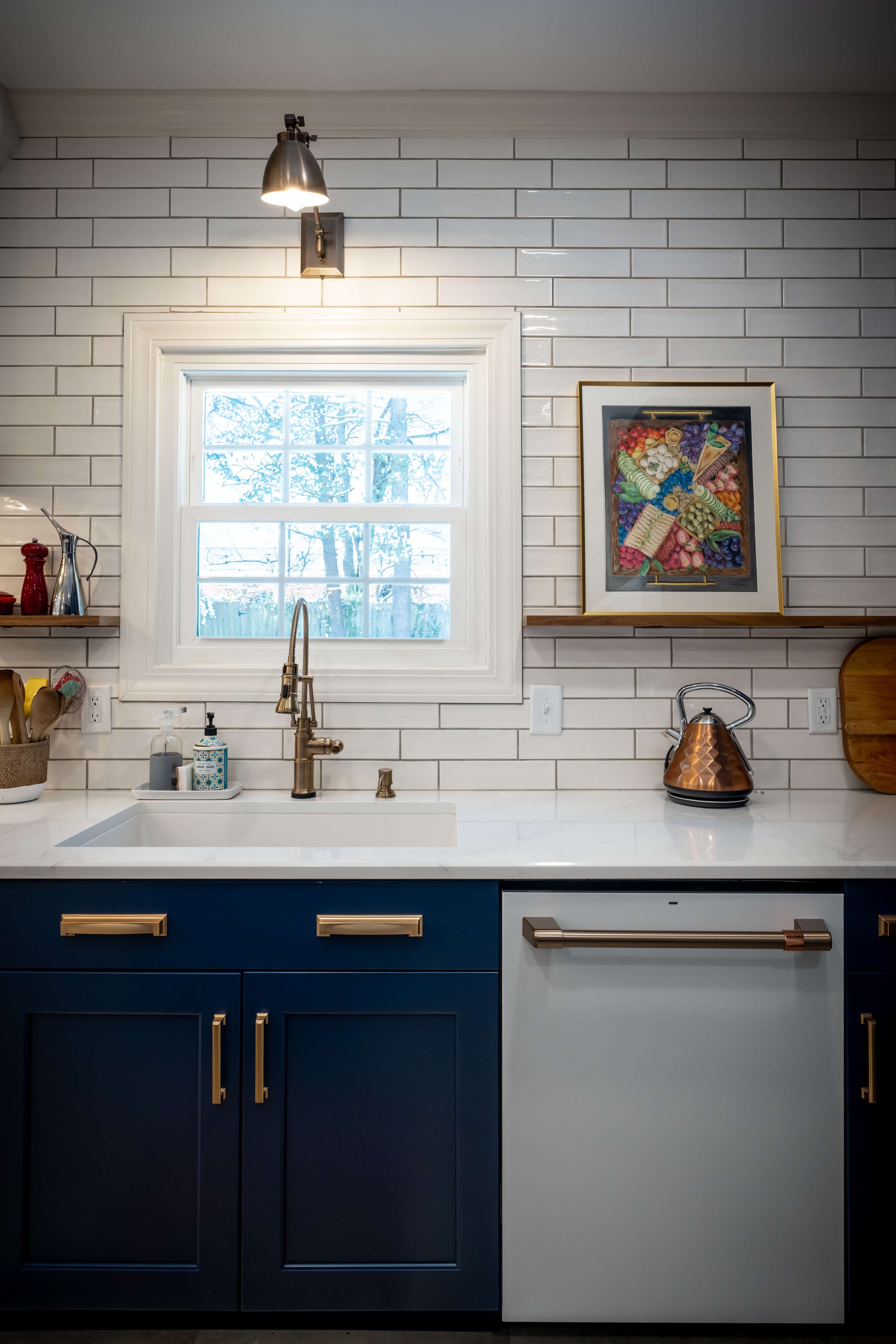 Modern kitchen sink with brass faucet, navy cabinets, white subway tile backsplash, window, kettle, and framed artwork.