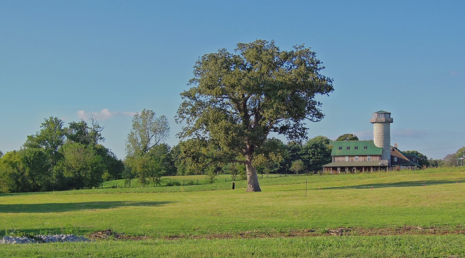 A scenic countryside landscape featuring a large green field, a prominent tree in the foreground, and a farmhouse with a tower in the background under a clear blue sky.
