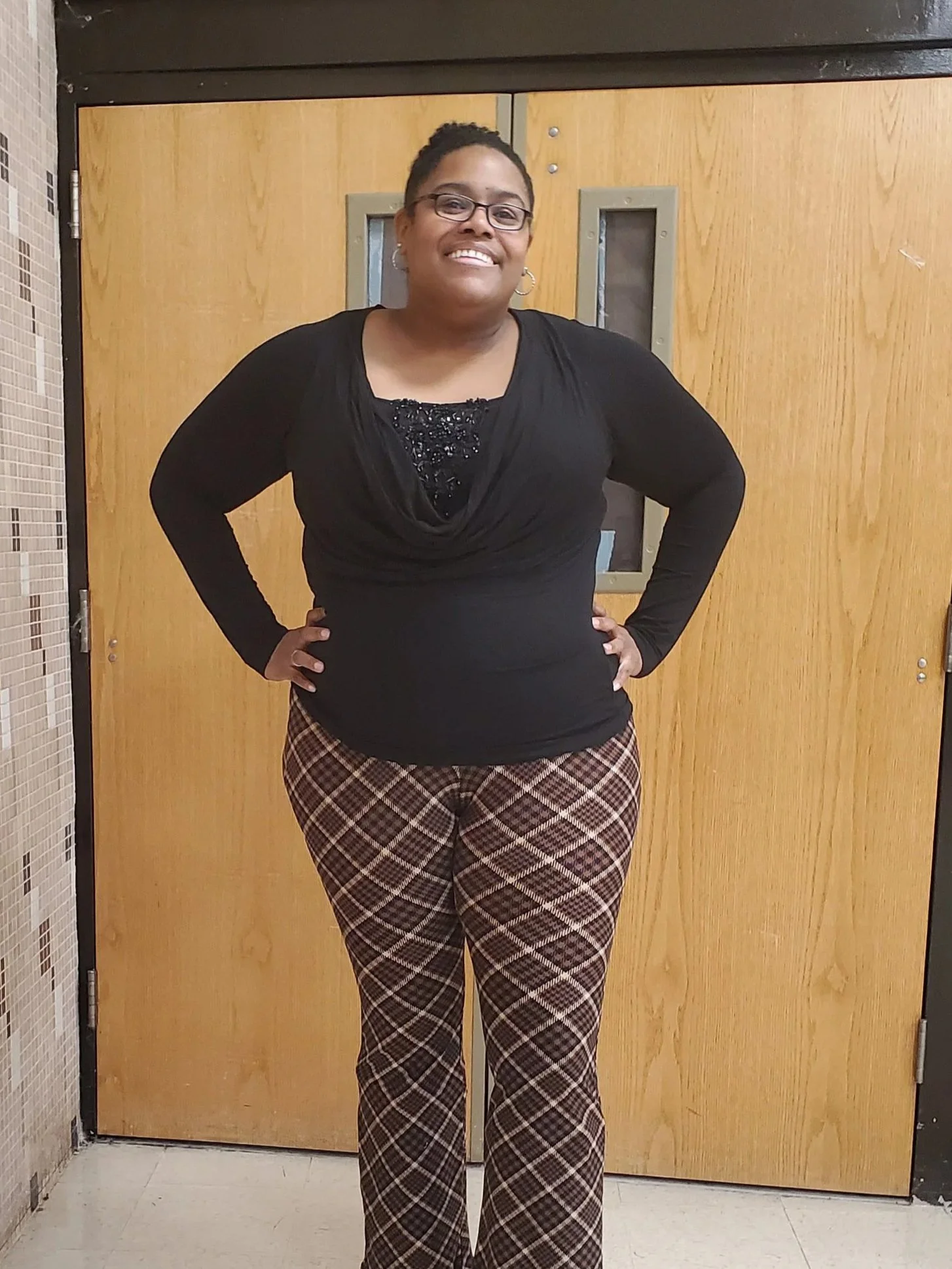 Chelcy is a black woman smiling at the camera with her hands on her hips. She wear glasses, and her hair is pulled back into a bun. 