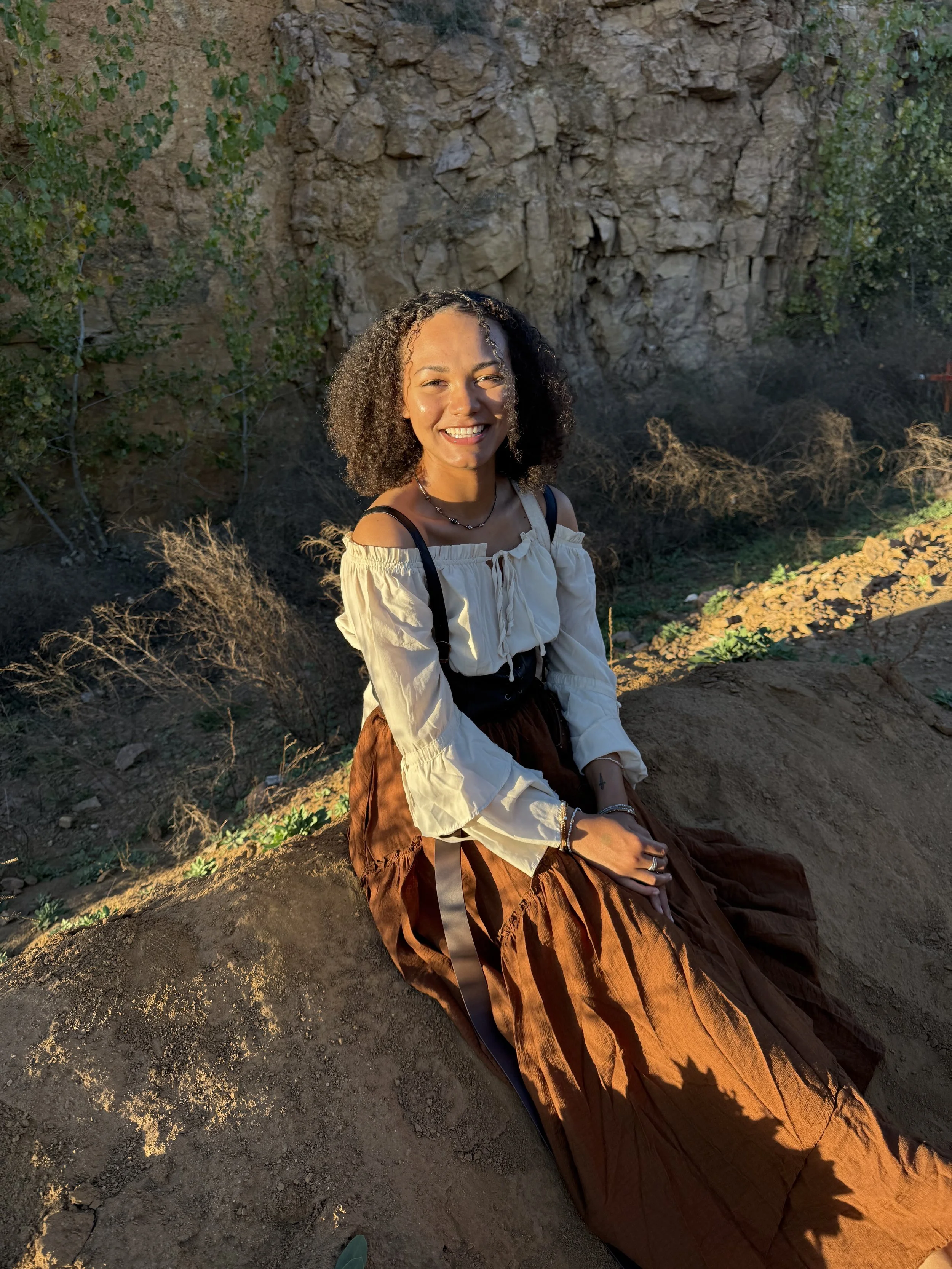 The sun is shining on the left side of Shykala's face as she sits outside. She is wearing a brown and white outfit, her hair is curly and pulled away from her face.
