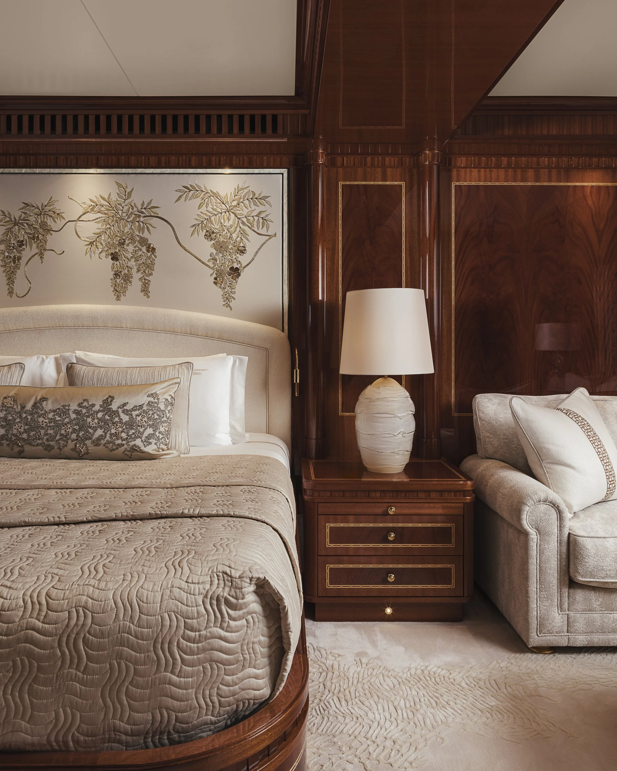 A bedroom with a bed featuring white pillows and a beige quilt, a wooden nightstand with drawers, a white table lamp, a beige armchair with a pillow, a wood-paneled wall, and a decorative mural of branches with leaves above the headboard.