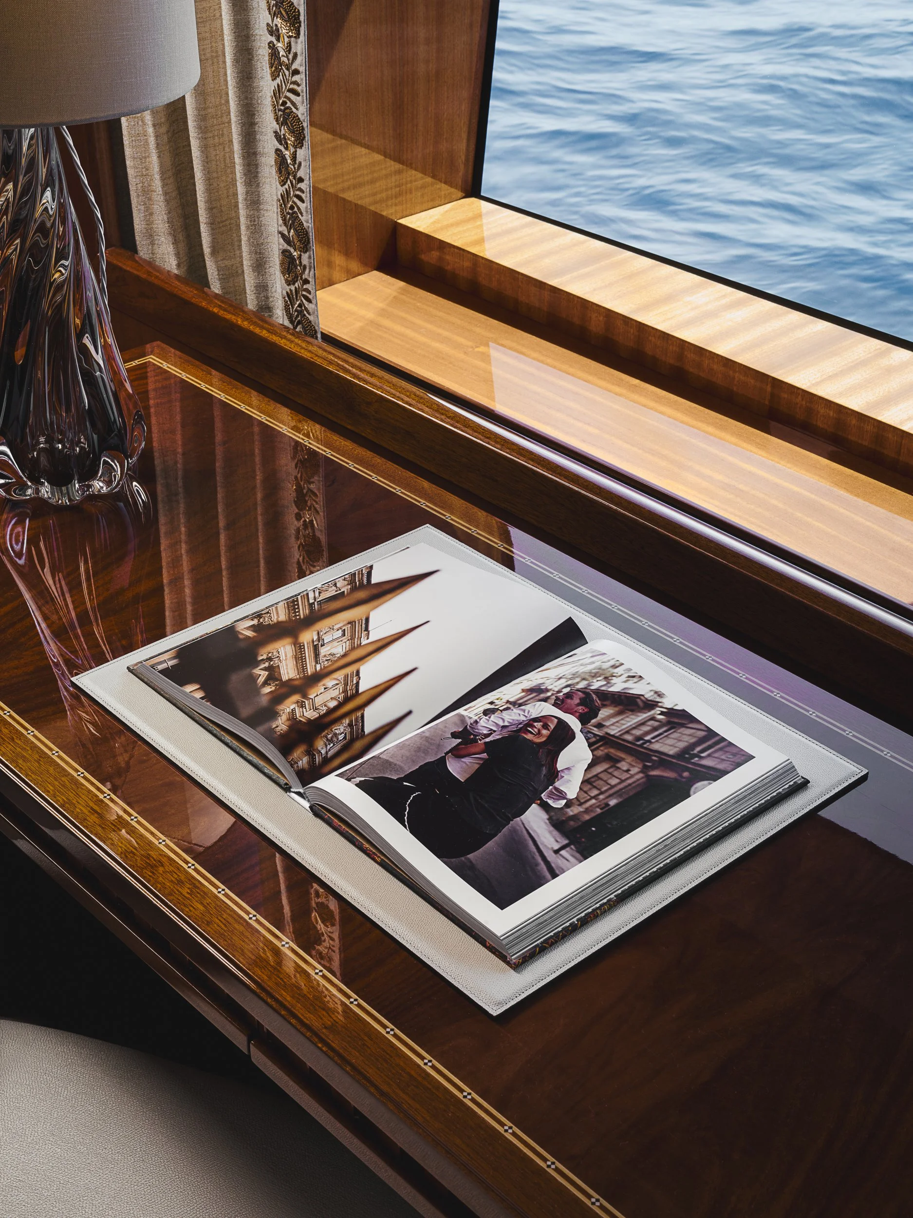 An open photo album on a polished wooden table with a glass of water nearby, displaying images of a smiling woman and a man in a historic city street scene with arches.
