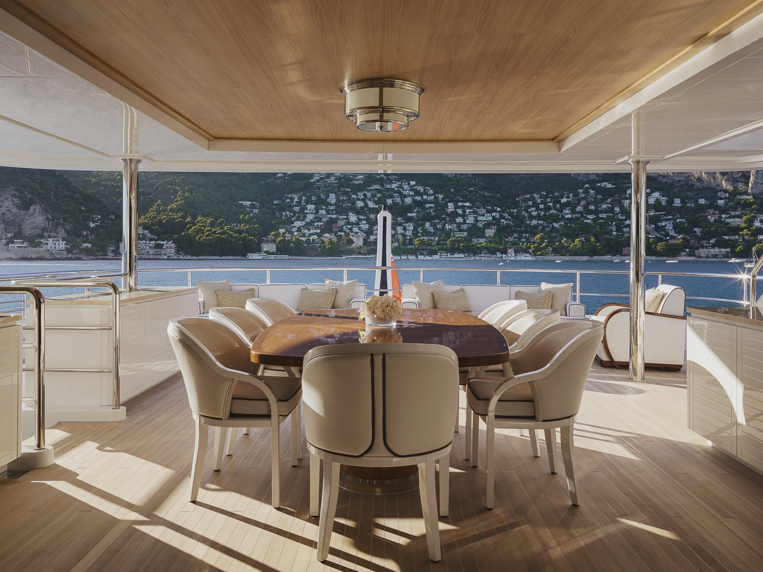 View from a yacht showing a dining area with a wooden table and white chairs, overlooking a body of water and a hillside with houses.