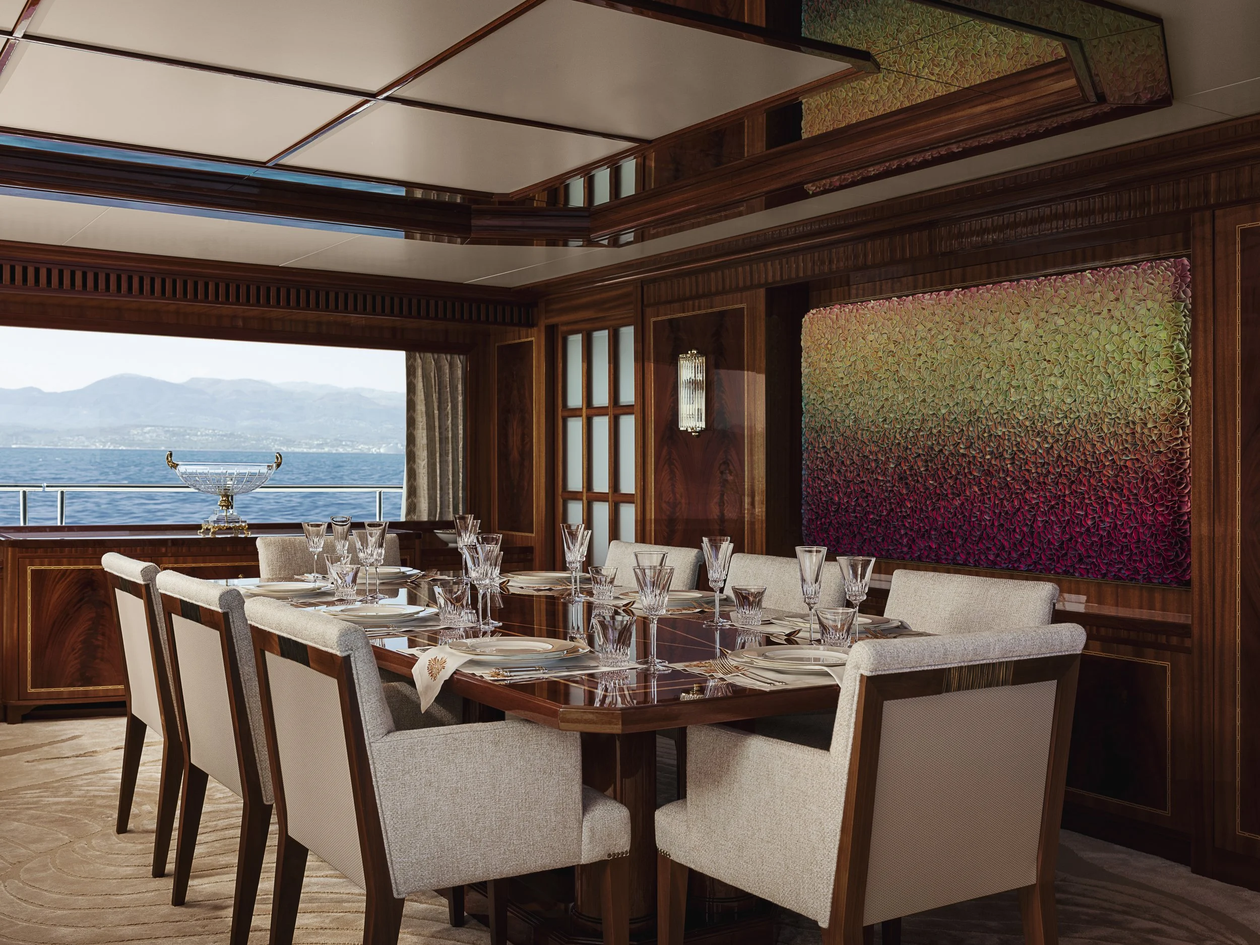 Elegant dining room with a large wooden table set with glasses and plates, beige upholstered chairs, large colorful wall art, and views of water and mountains outside through windows.