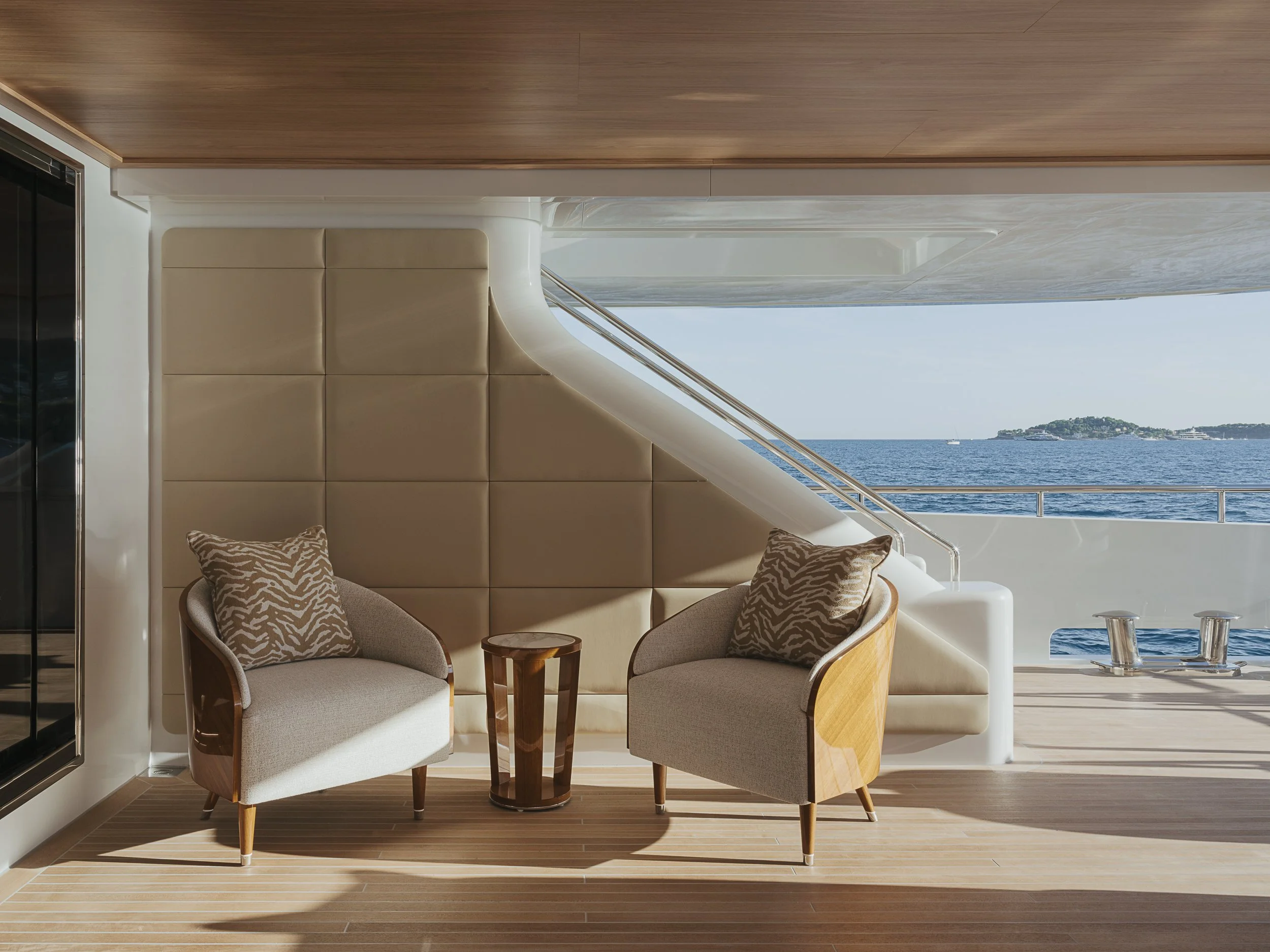 Interior of a yacht with two beige armchairs with patterned pillows, a small wooden table, and a view of the ocean and distant land through a large window.