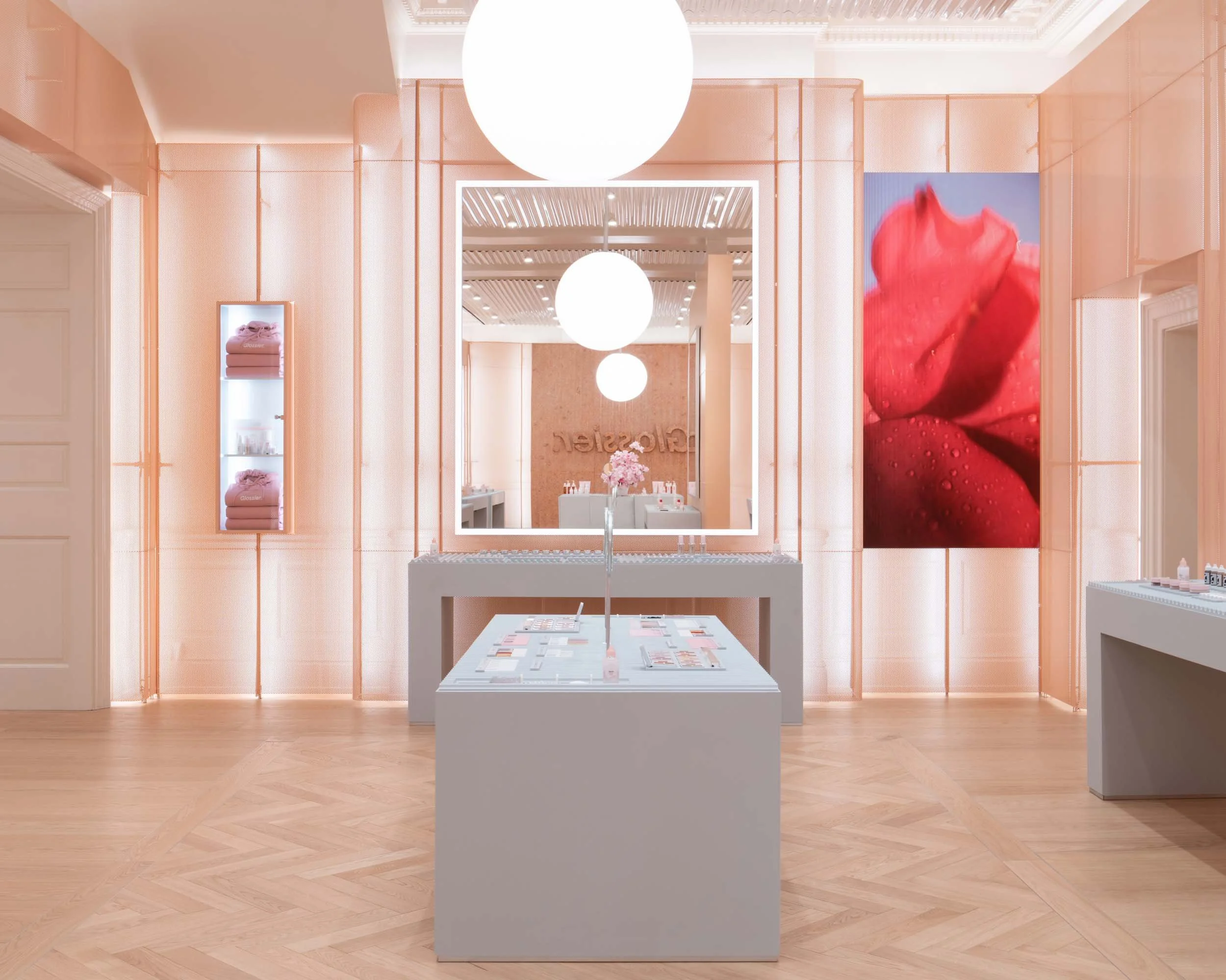 A modern, minimalist retail store with soft pink walls, large white spherical ceiling lights, and light wooden flooring. The space displays products on white tables and shelves, with a large mirror and a digital screen showing a close-up of pink rose petals with water droplets.