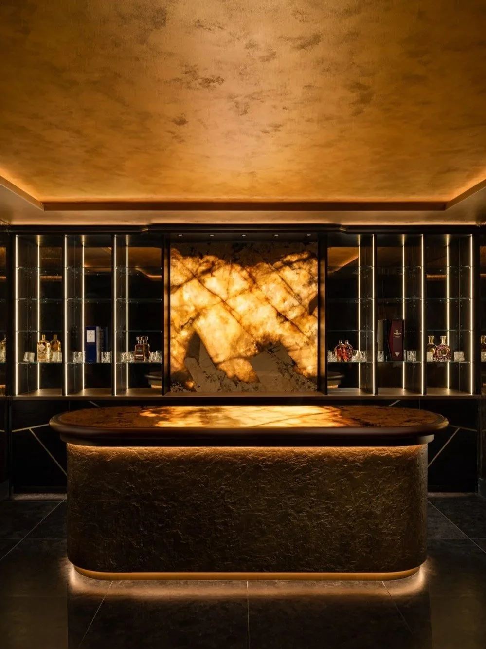 Luxury bar interior featuring a backlit onyx counter, ambient golden lighting, and sleek glass shelving with premium spirits, showcasing high-end hospitality design and architectural photography.