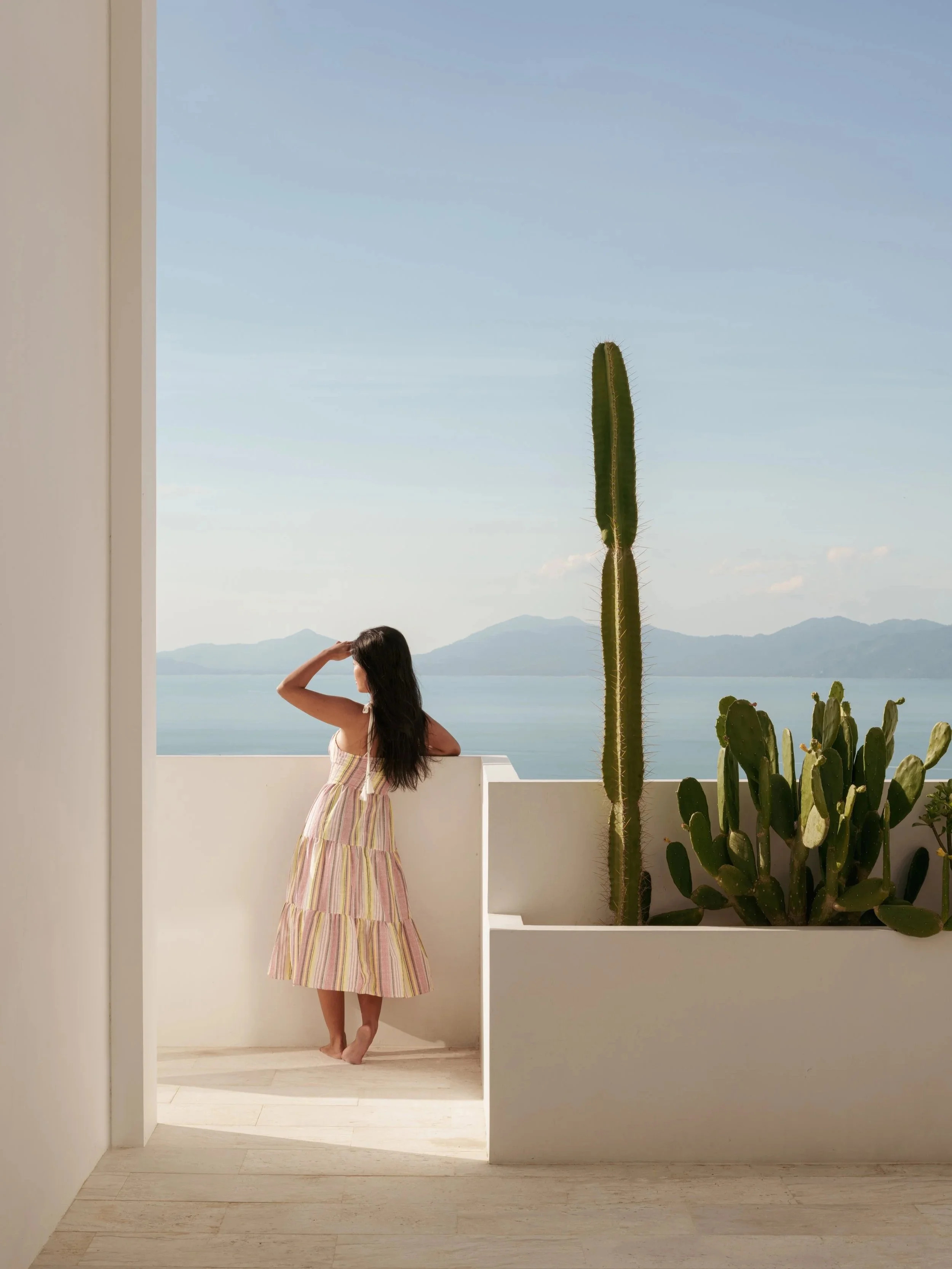 Minimalist coastal terrace with white modern architecture, lush cacti, and ocean views, as a woman gazes at the horizon, highlighting luxury hospitality and architectural design photography.
