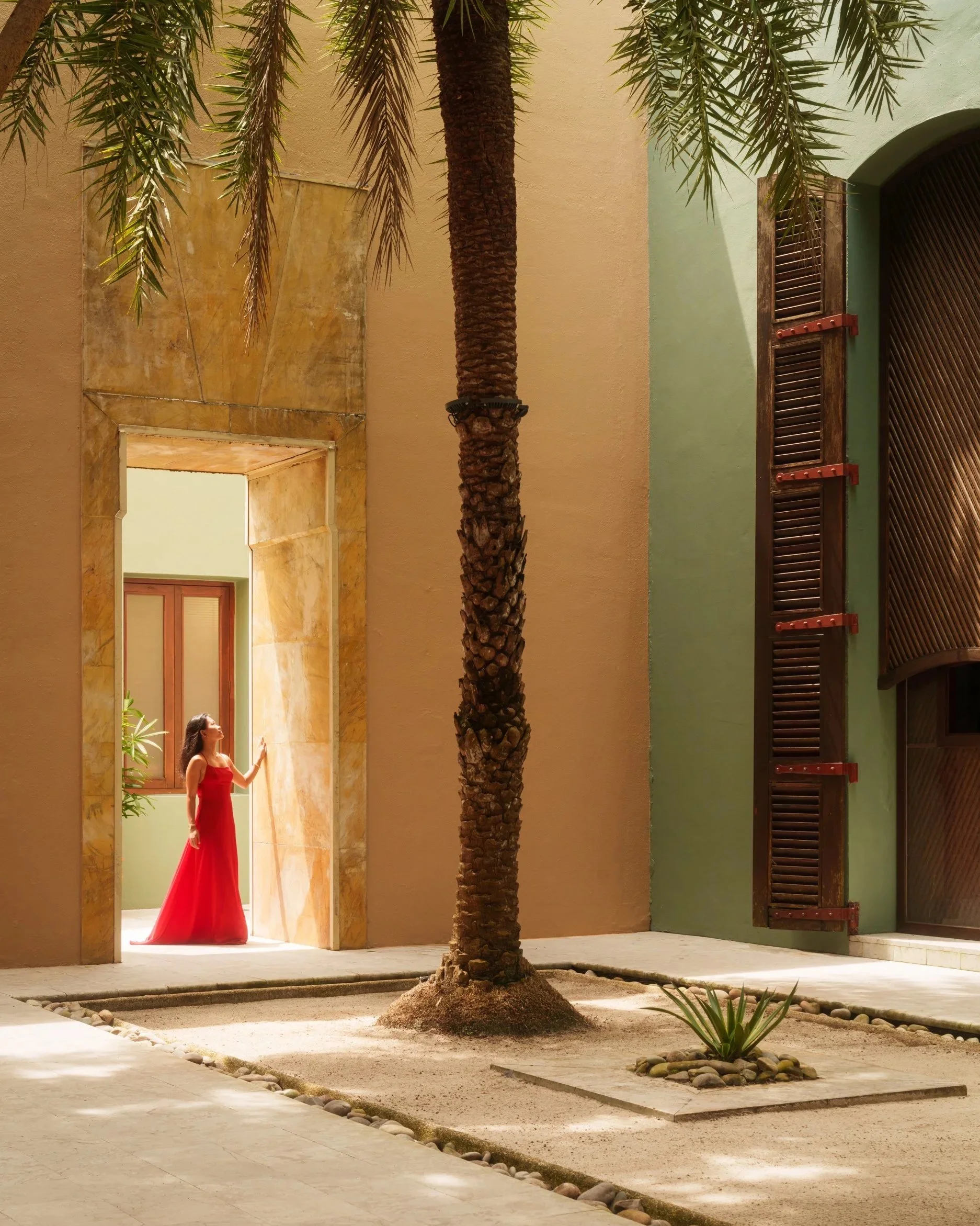 Serene courtyard with Mediterranean-inspired architecture, warm-toned walls, and tropical greenery, featuring a woman in a red dress, showcasing luxury hospitality and architectural photography.
