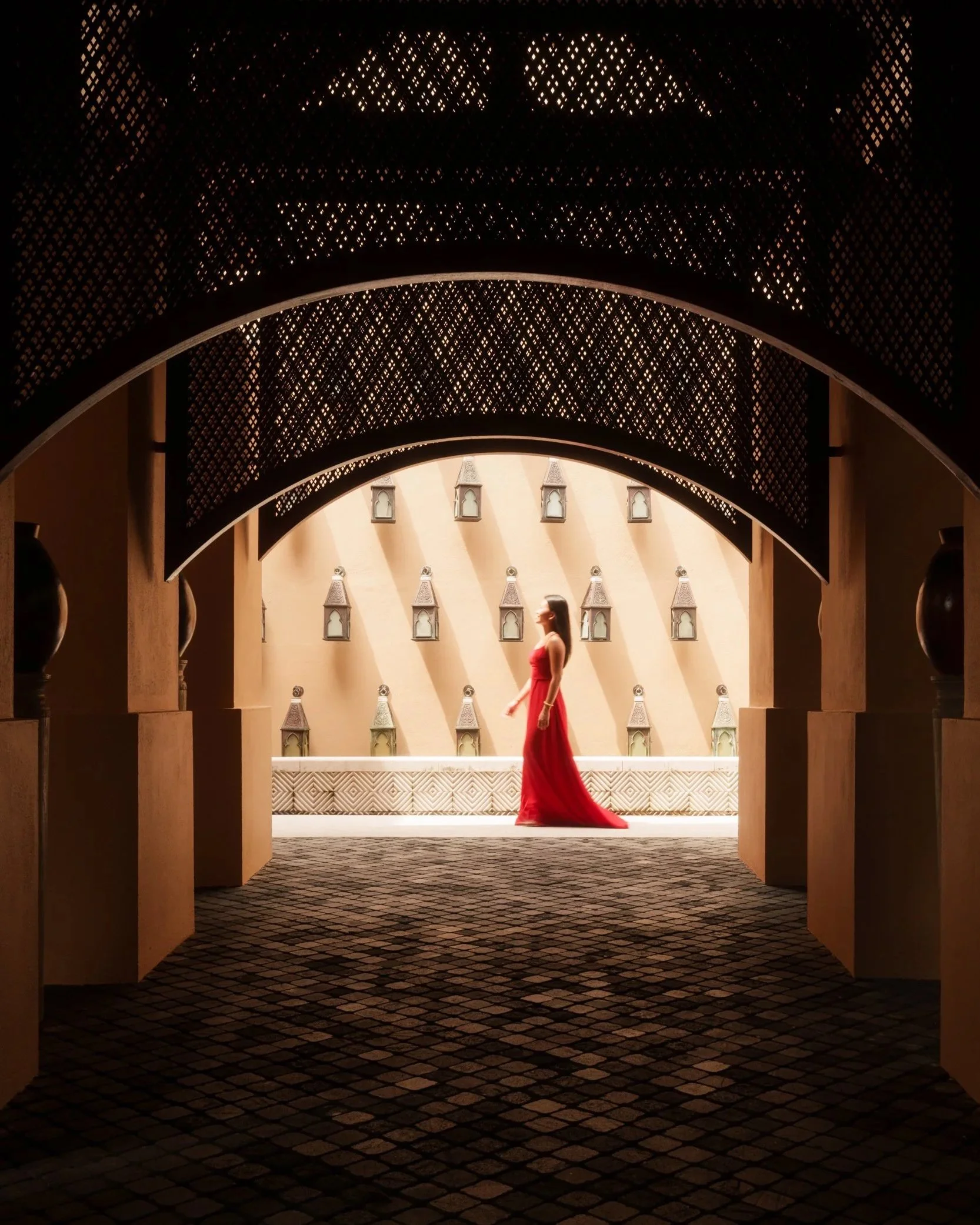 Architectural photography of an arched corridor with intricate latticework, featuring a woman in a red dress walking through a sunlit courtyard.