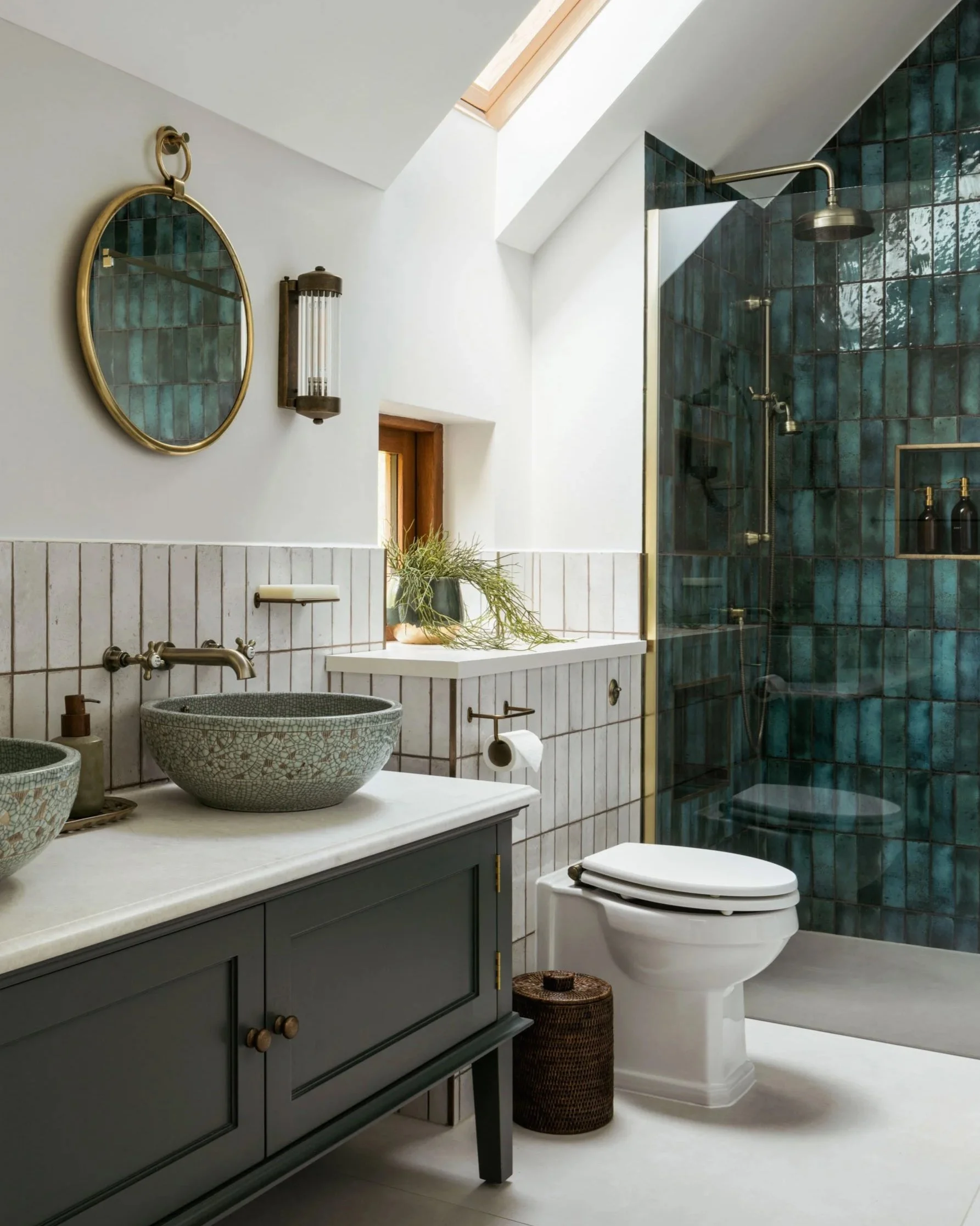 Elegant bathroom with a deep green vanity, artisanal vessel sinks, and textured blue shower tiles. The skylight and warm brass accents enhance the space with natural light and character.