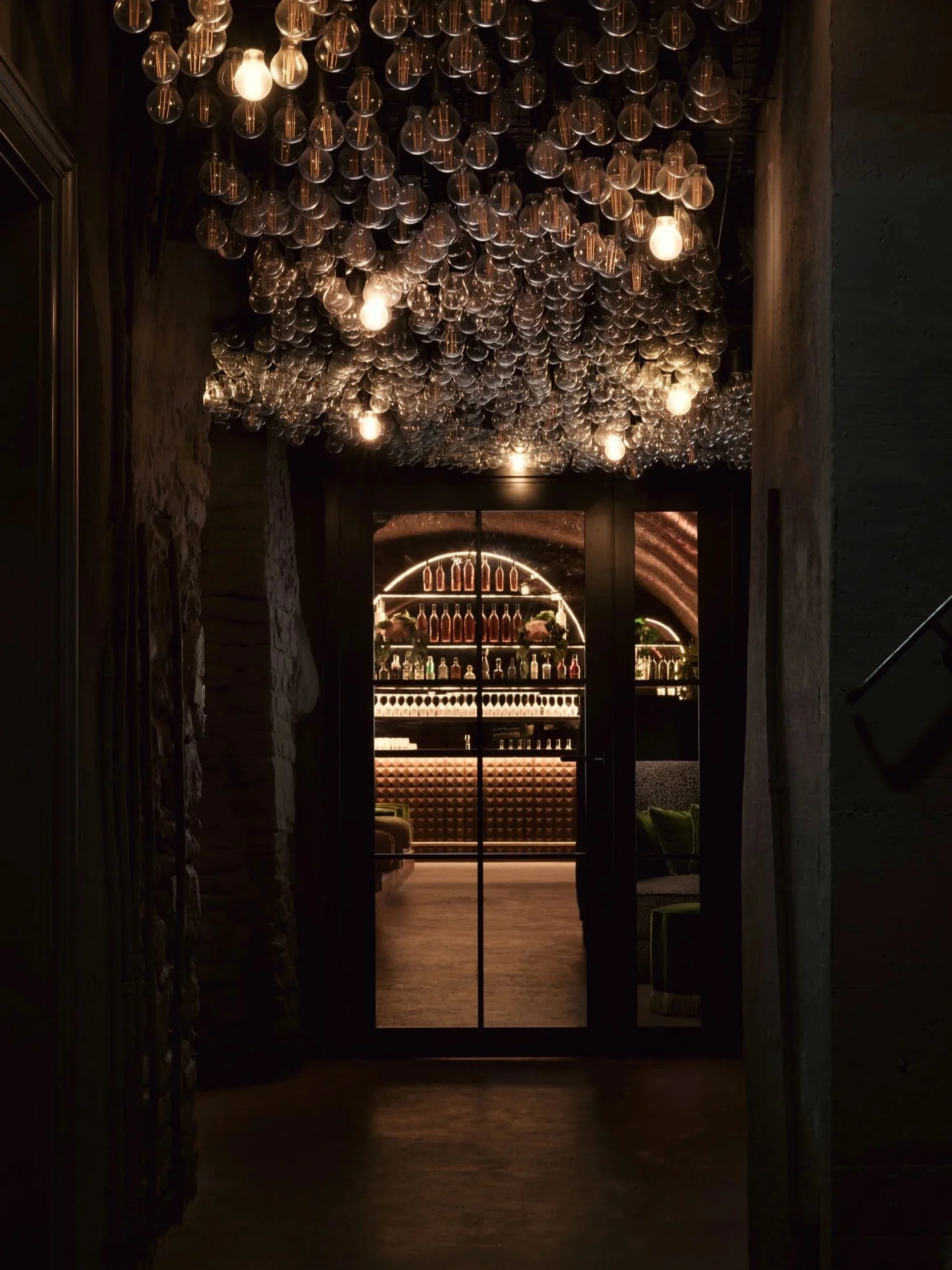 Atmospheric entrance to an underground bar in London, featuring a ceiling installation of hanging light bulbs and a softly illuminated arched back bar.