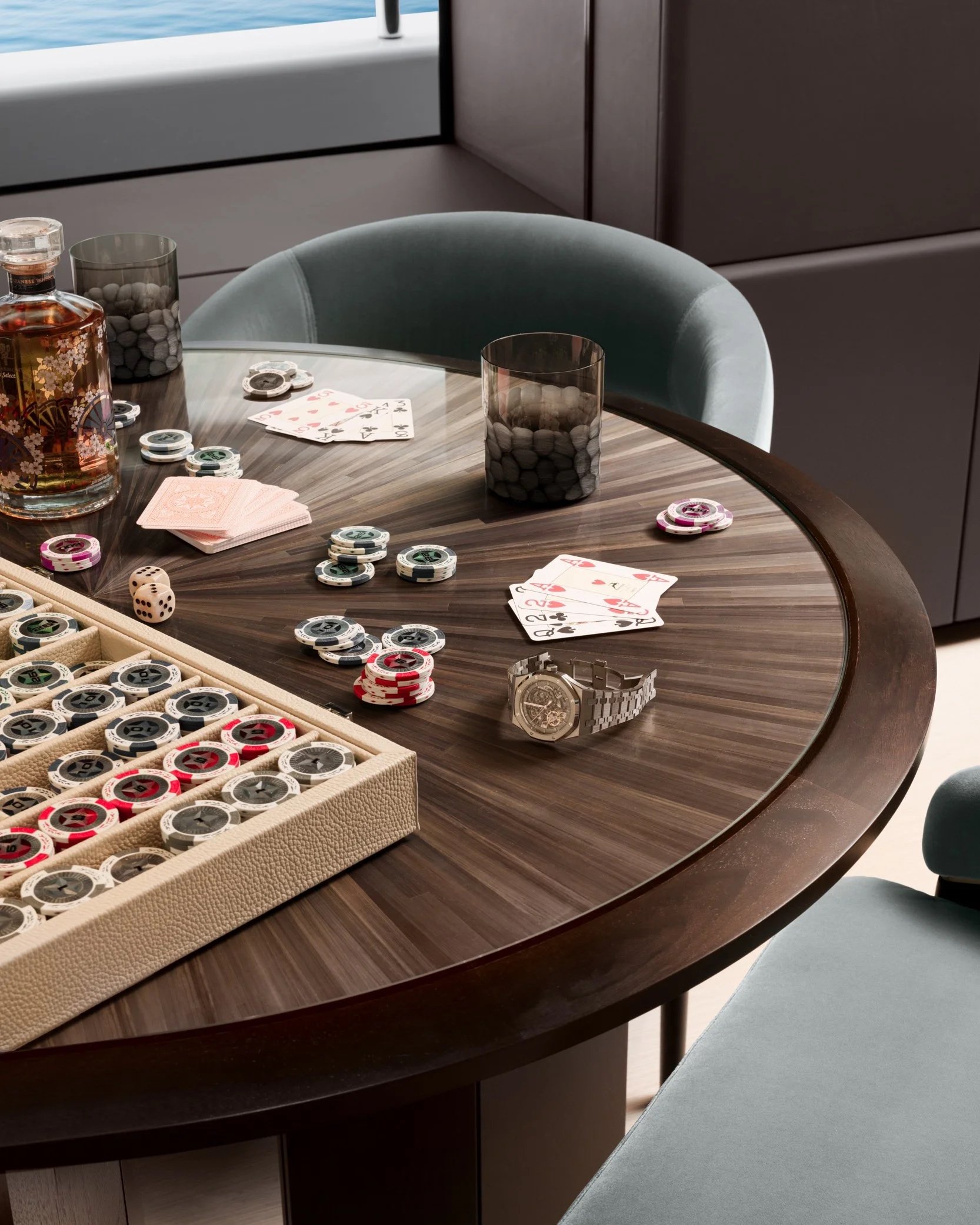Luxury poker table setup featuring a dark wood circular table, plush velvet chairs, poker chips, playing cards, and a whiskey decanter, showcasing high-end interior design and hospitality photography.