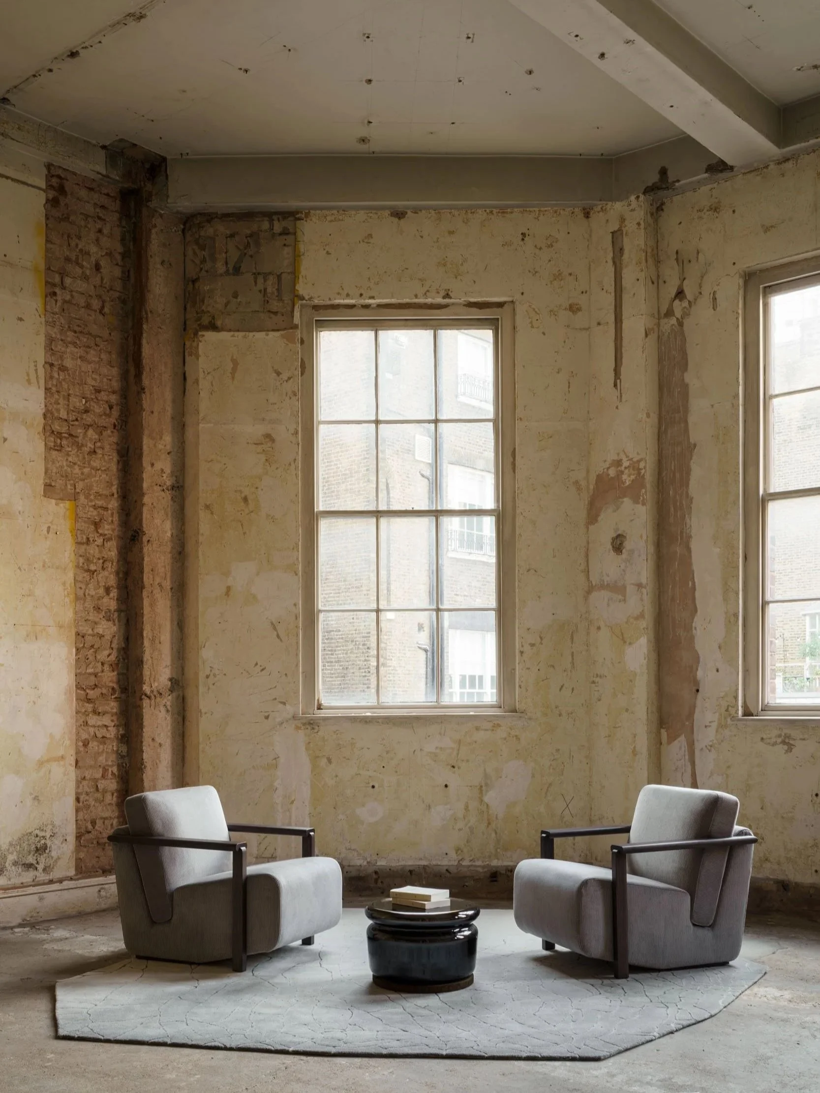Raw and textured interior in London featuring exposed plaster walls, industrial windows, and a minimalist seating arrangement with sculptural armchairs and a central coffee table.