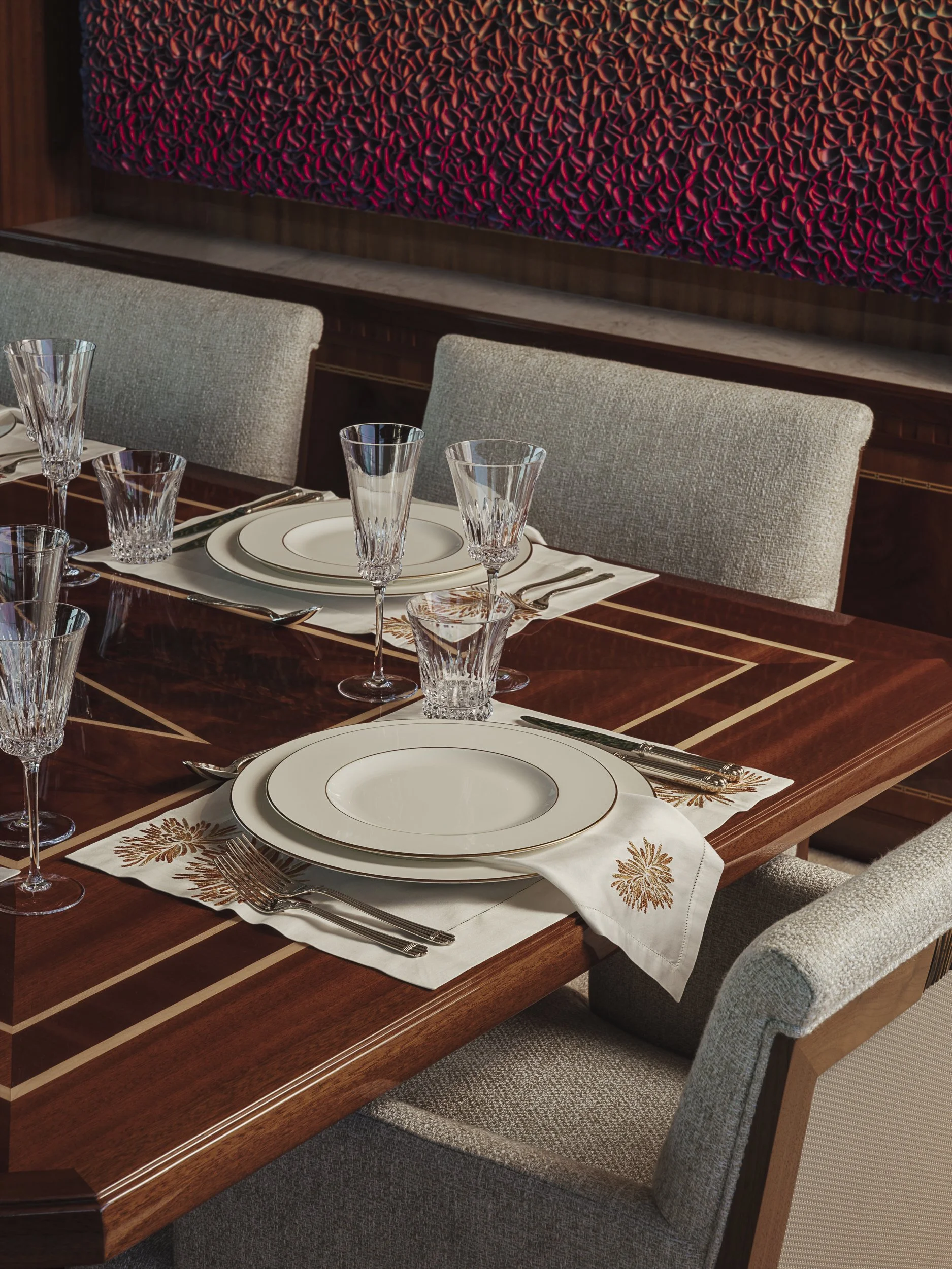 Elegant dining table set with white plates, crystal glasses, and silverware on a white and gold embroidered tablecloth. Surrounding chairs are upholstered in light gray fabric. The background features a textured red and black art piece on a wooden wa