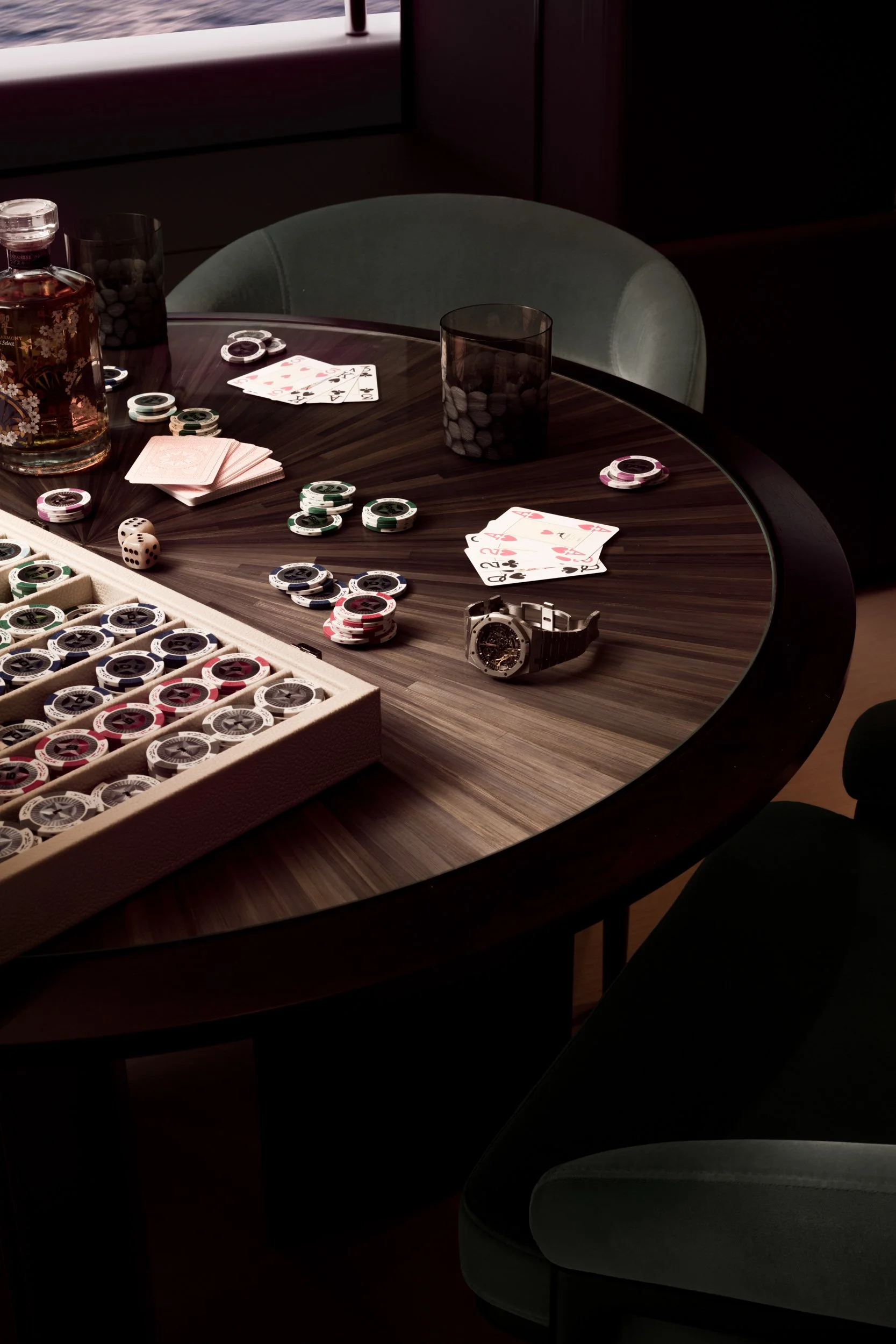 A poker game setup on a wooden table with chips, playing cards, a deck, a watch, and a bottle of liquor. Two empty glasses and a green upholstered chair are also visible.